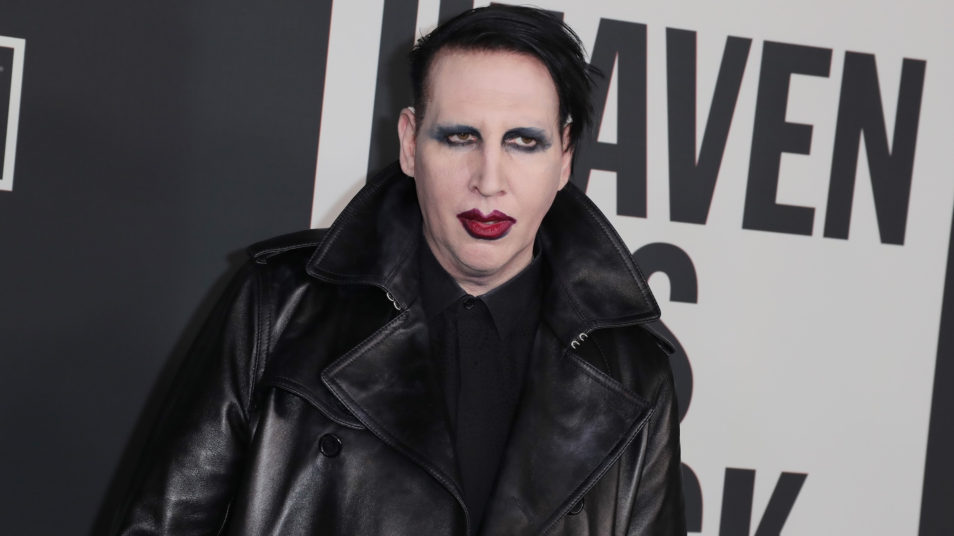 Marilyn Manson in a black leather coat with dark makeup, standing in front of a sign with partial text.