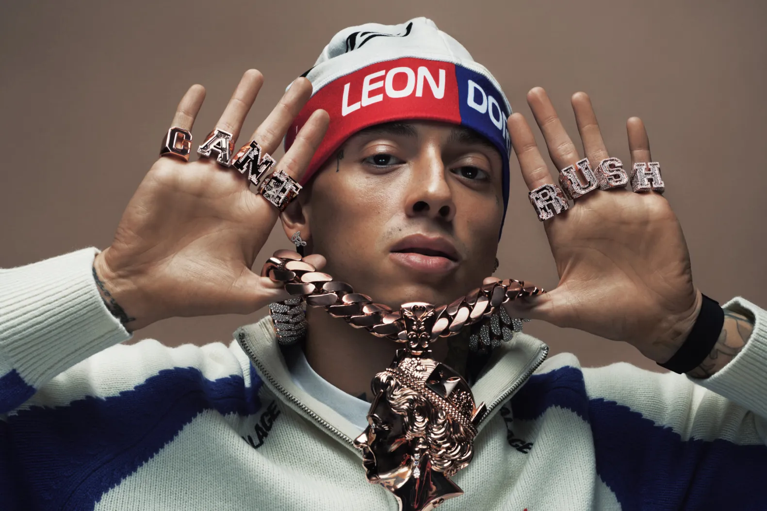 Central Cee wearing a "LEON DORE" beanie, large chain necklace, and rings spelling "CAN'T RUSH." Hands are raised near face, casual attire.