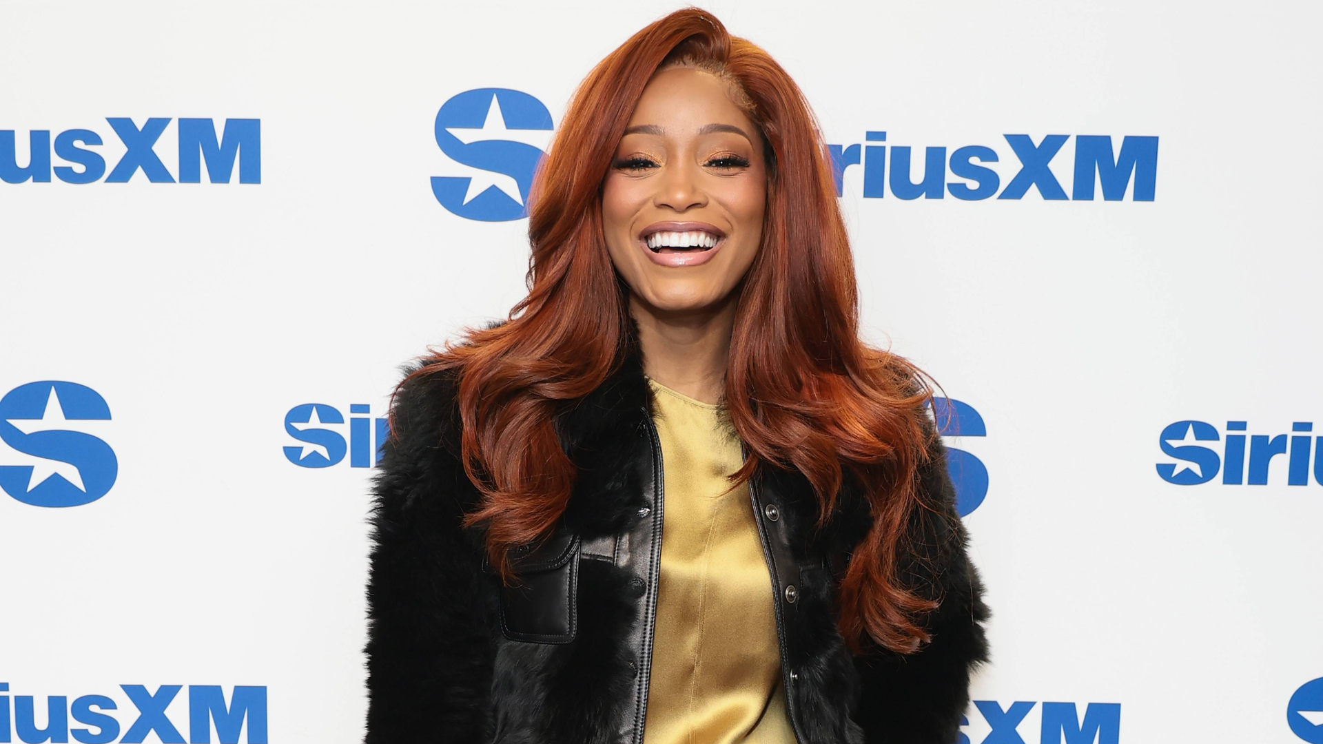Keke Palmer smiling in front of a SiriusXM backdrop, wearing a black jacket over a gold top.