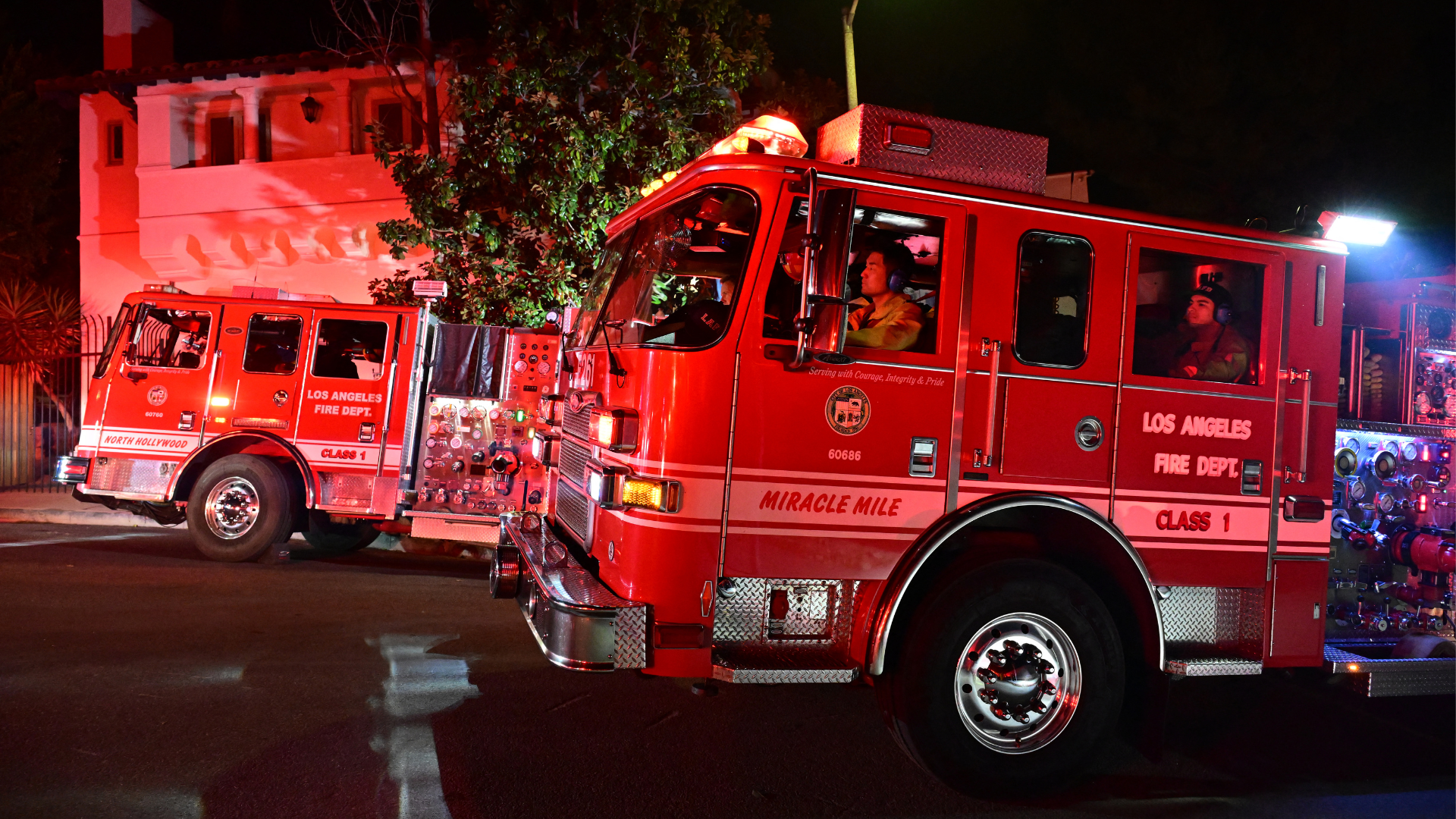 Two red Los Angeles Fire Department trucks with firefighters inside, parked on a street at night.