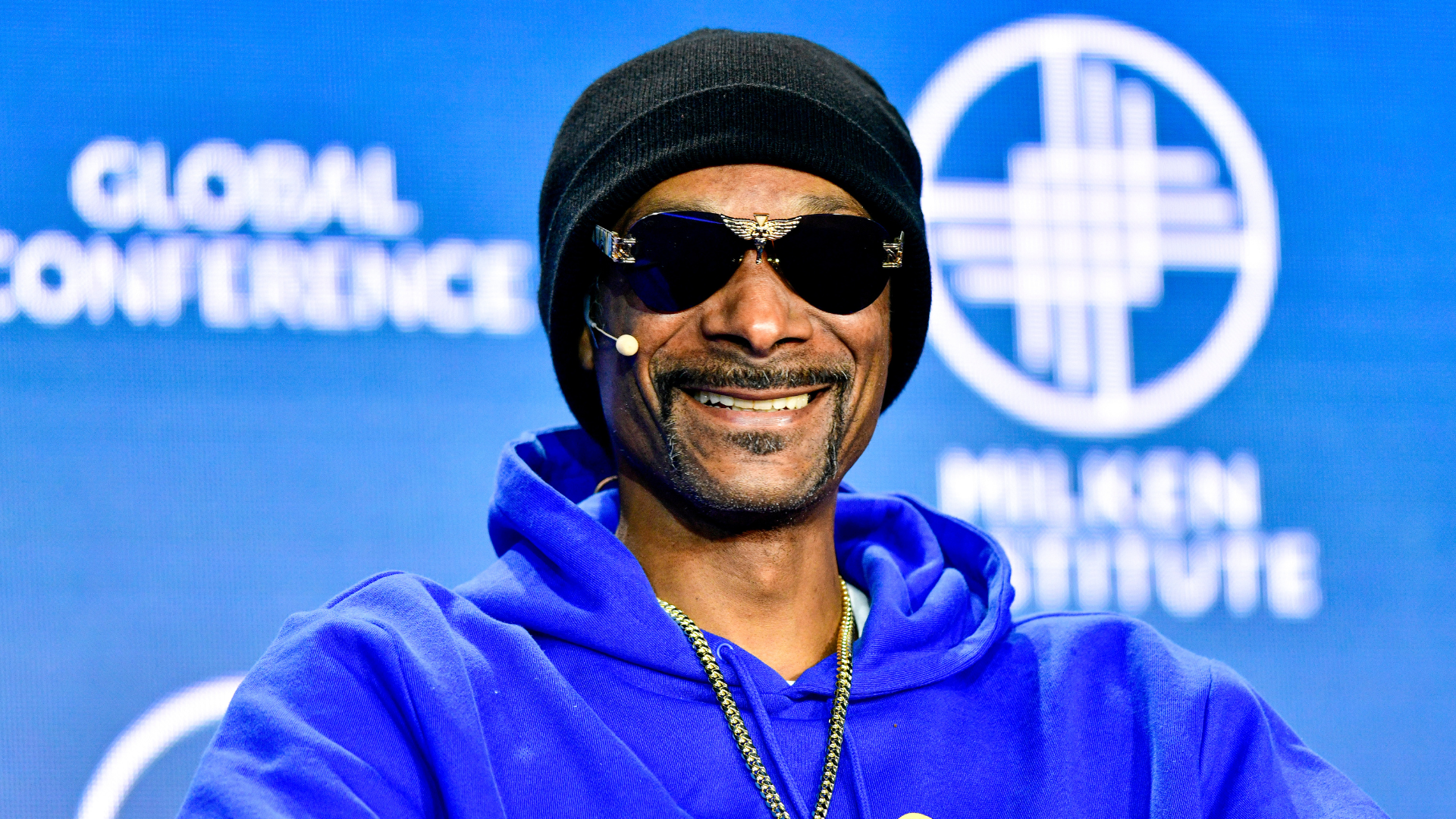 Snoop Dogg wearing sunglasses and a blue hoodie, smiling at a conference with a blue background.