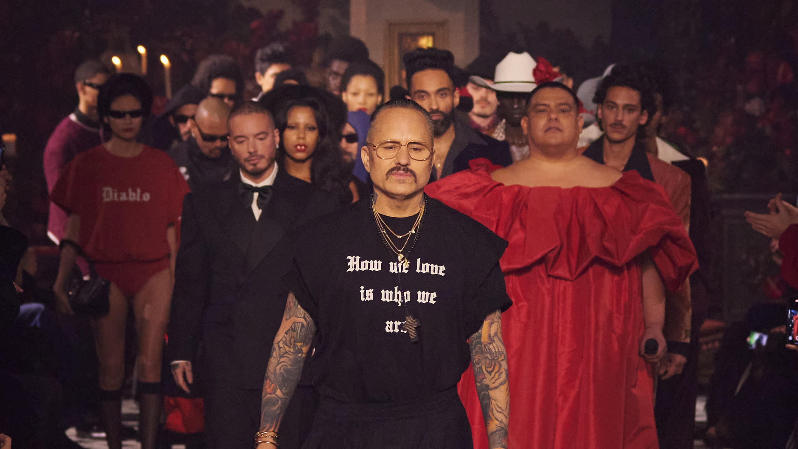 A diverse group of models, including a person in a black shirt with tattoos and another in a red dress, walk down a dimly lit runway.