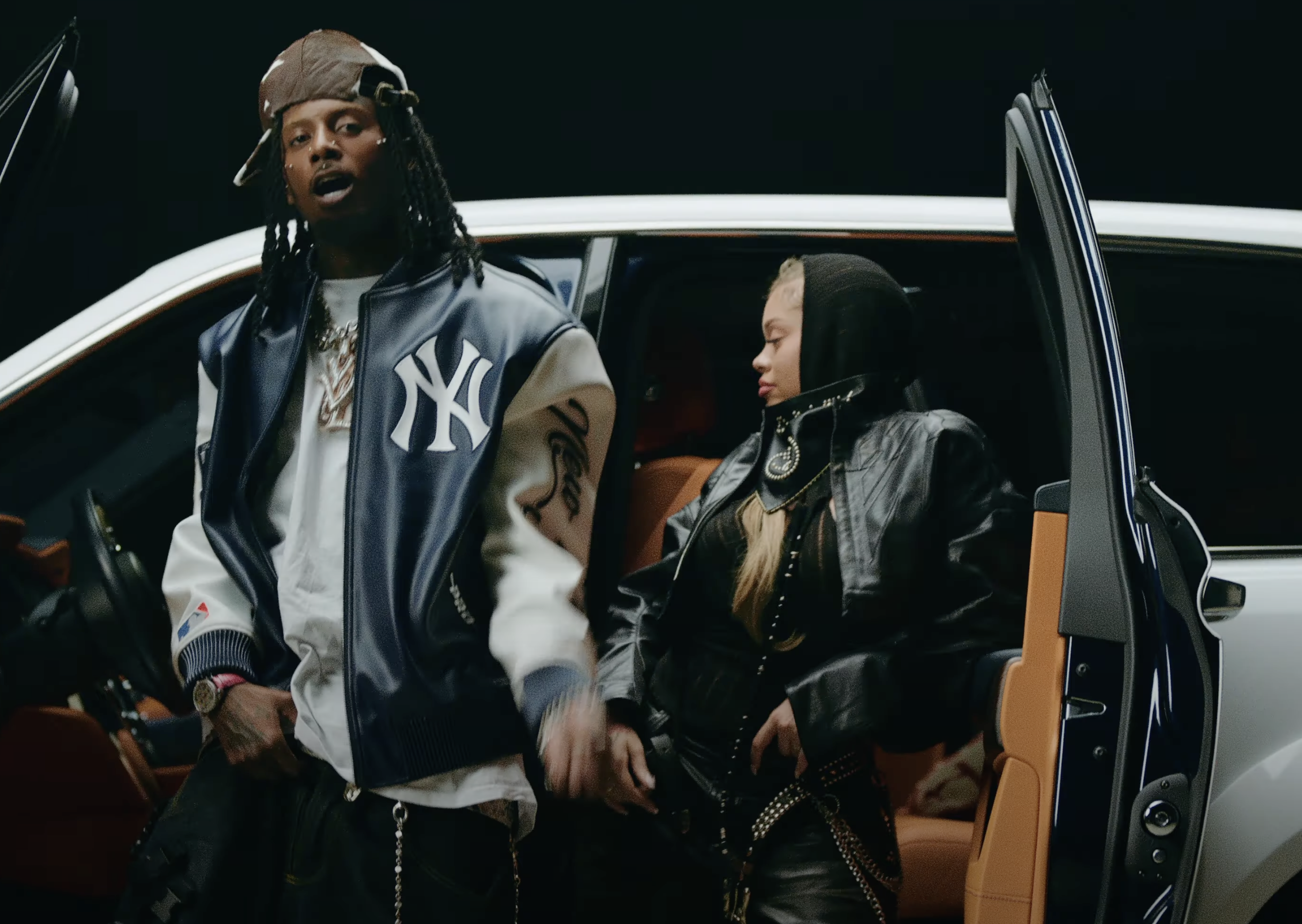 Two people stand by an open car door. One wears a New York Yankees jacket and cap, the other a black leather jacket with a hood.