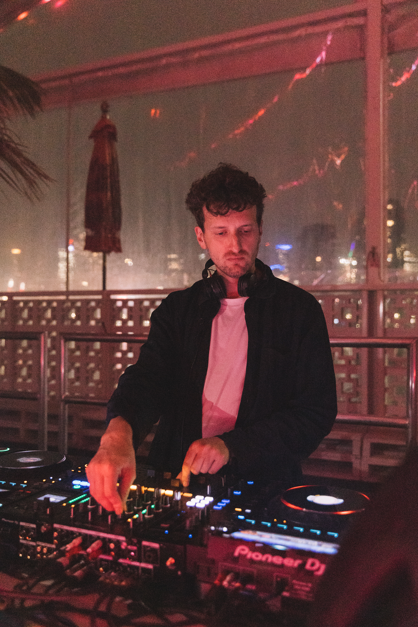 A DJ with curly hair is performing at a mixer with headphones around his neck in a dimly lit setting.