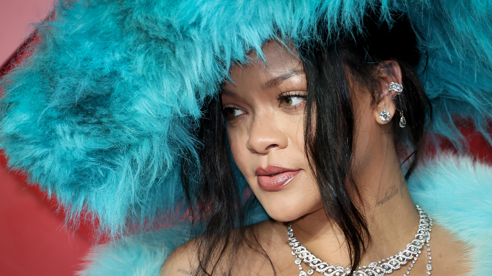 Rihanna wearing a large blue feathered hat and matching outfit, adorned with sparkling jewelry, looking to the side.