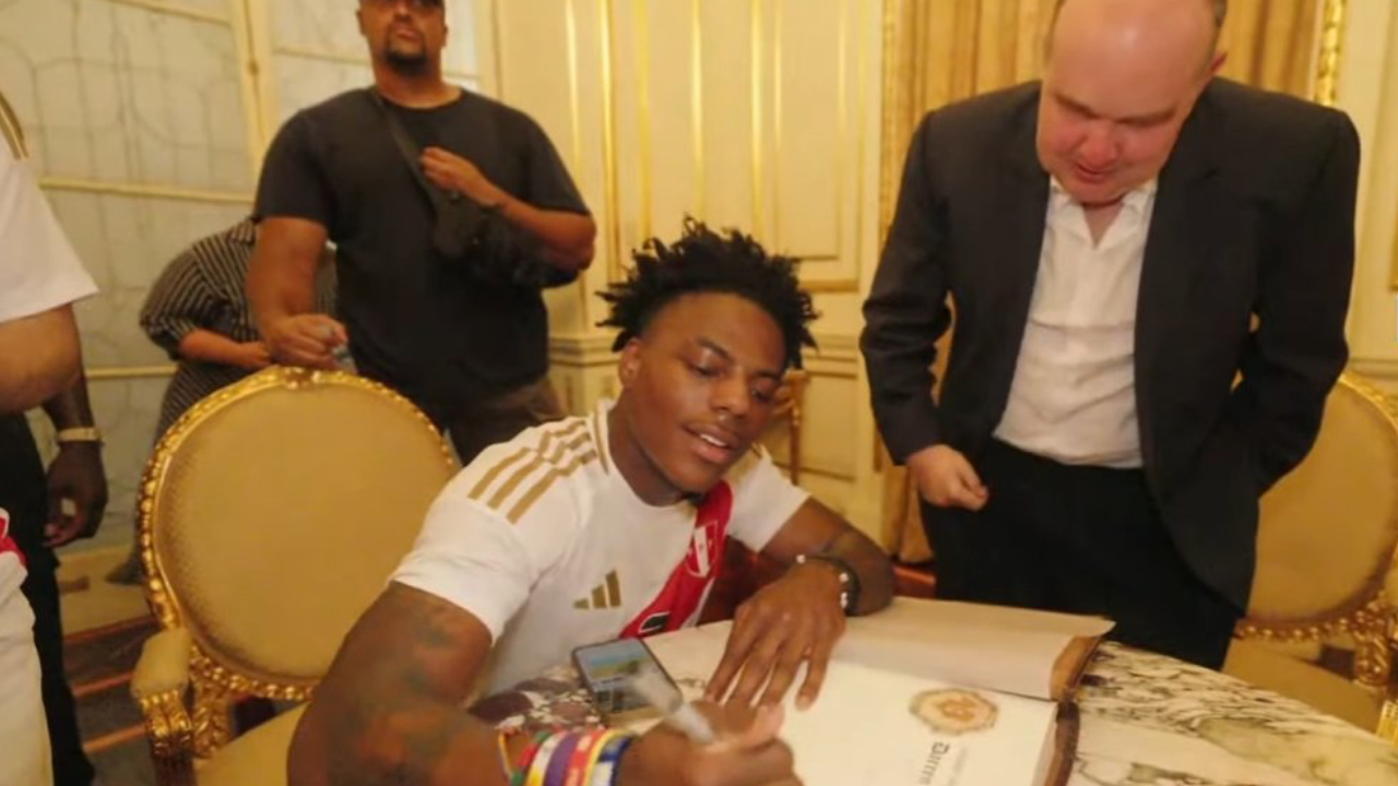 A man in a sports jersey signs a book at a table, surrounded by others in a room with ornate decor.