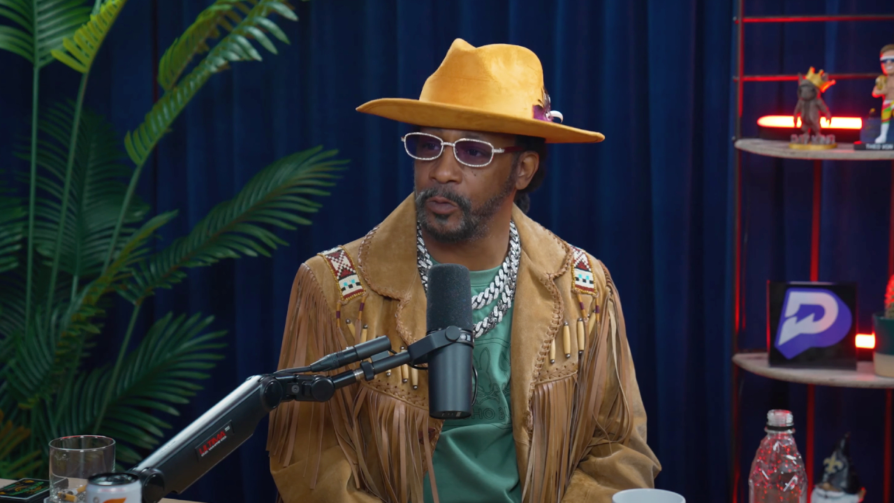 Katt Williams wearing a yellow hat, sunglasses, and a fringed jacket speaks into a microphone in a studio setting with plants and shelves.
