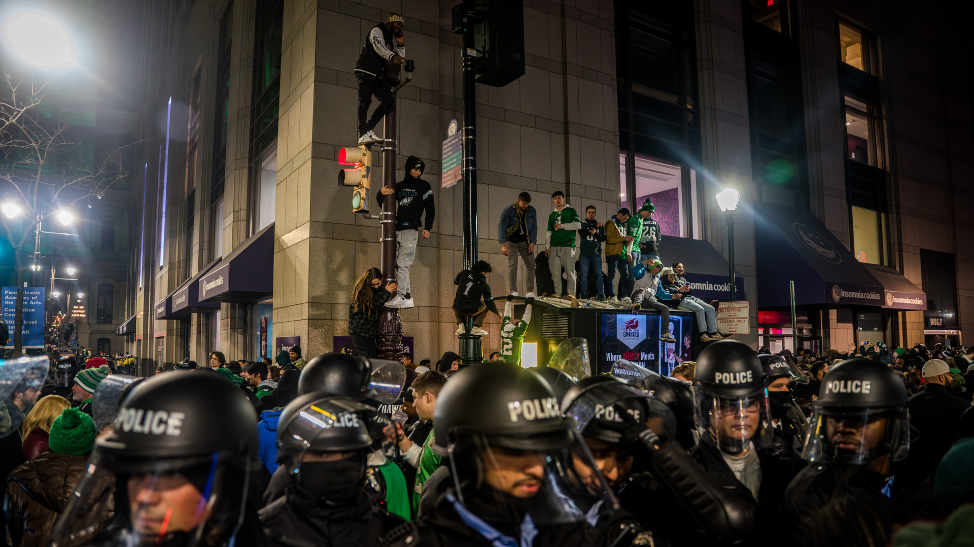 A crowd of people, some climbing a building, with police officers in riot gear in the foreground. It's nighttime in an urban setting.
