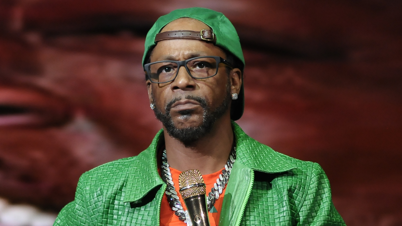 Katt Williams wearing a green jacket and cap, holding a microphone.
