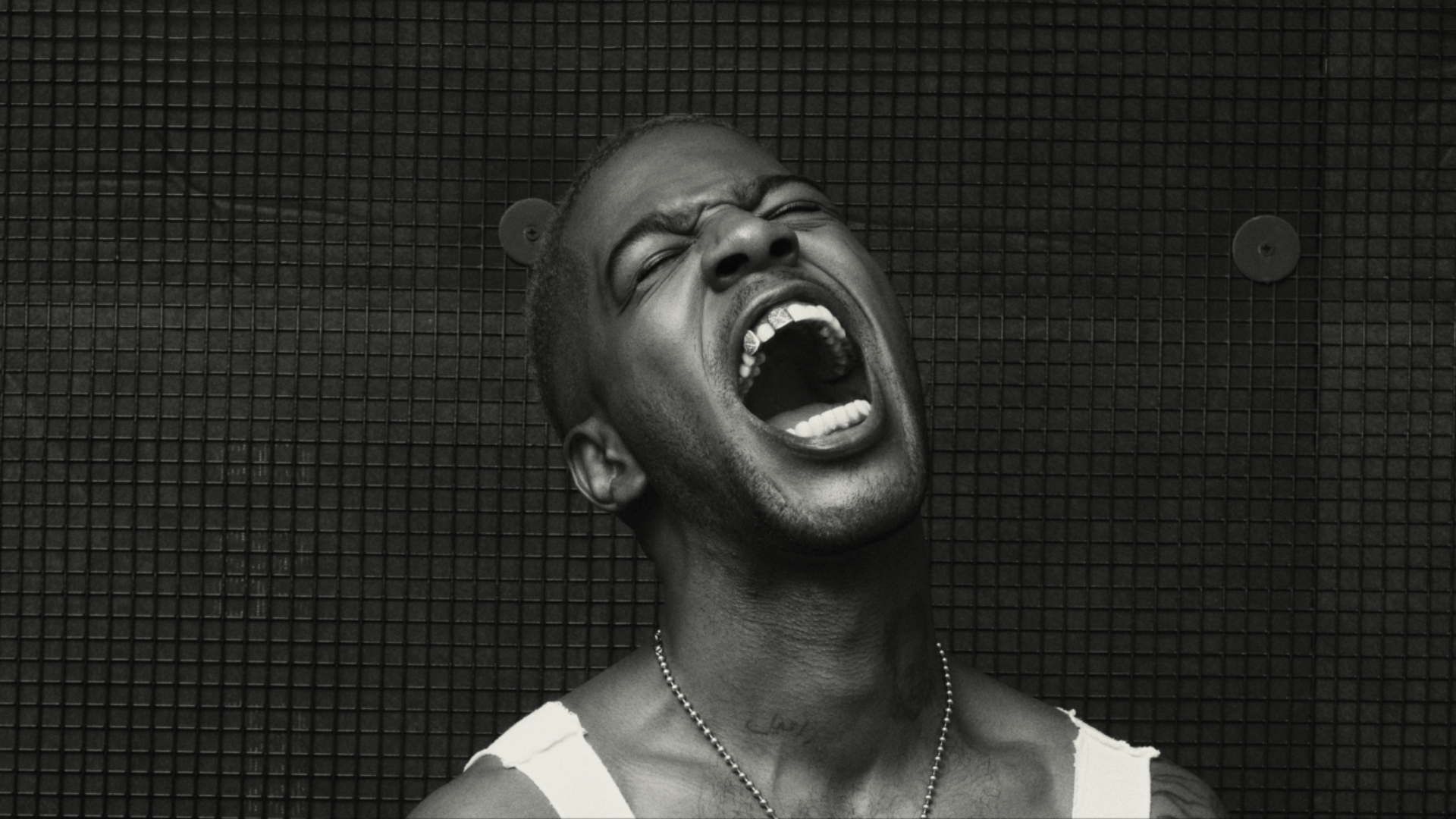 Kid Cudi in a tank top with an open mouth, expressing strong emotion against a dark, textured background.