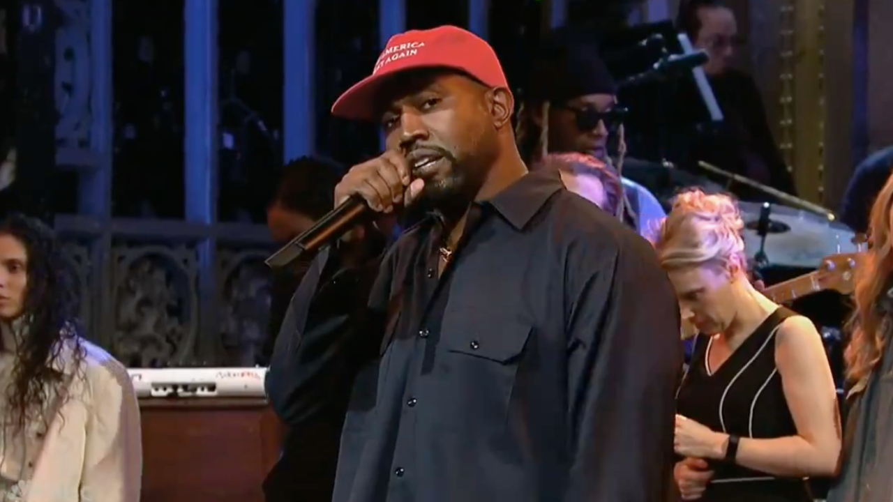 Ye wearing a red cap, performing on stage with a microphone, surrounded by other people and musical instruments.
