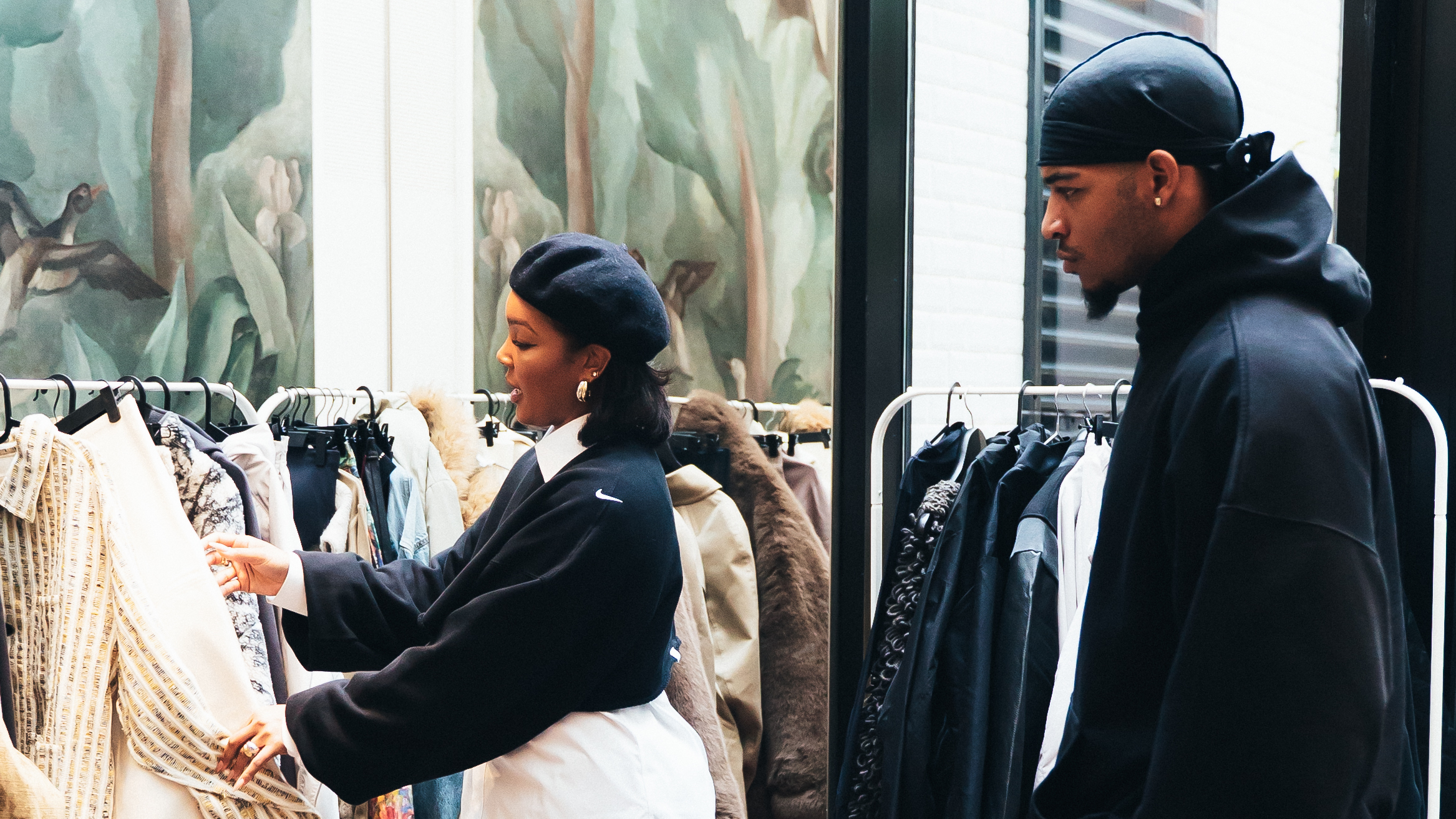 Two people are browsing through clothing racks in a store. The woman wears a beret and a black jacket, while the man wears a black hoodie and a durag.