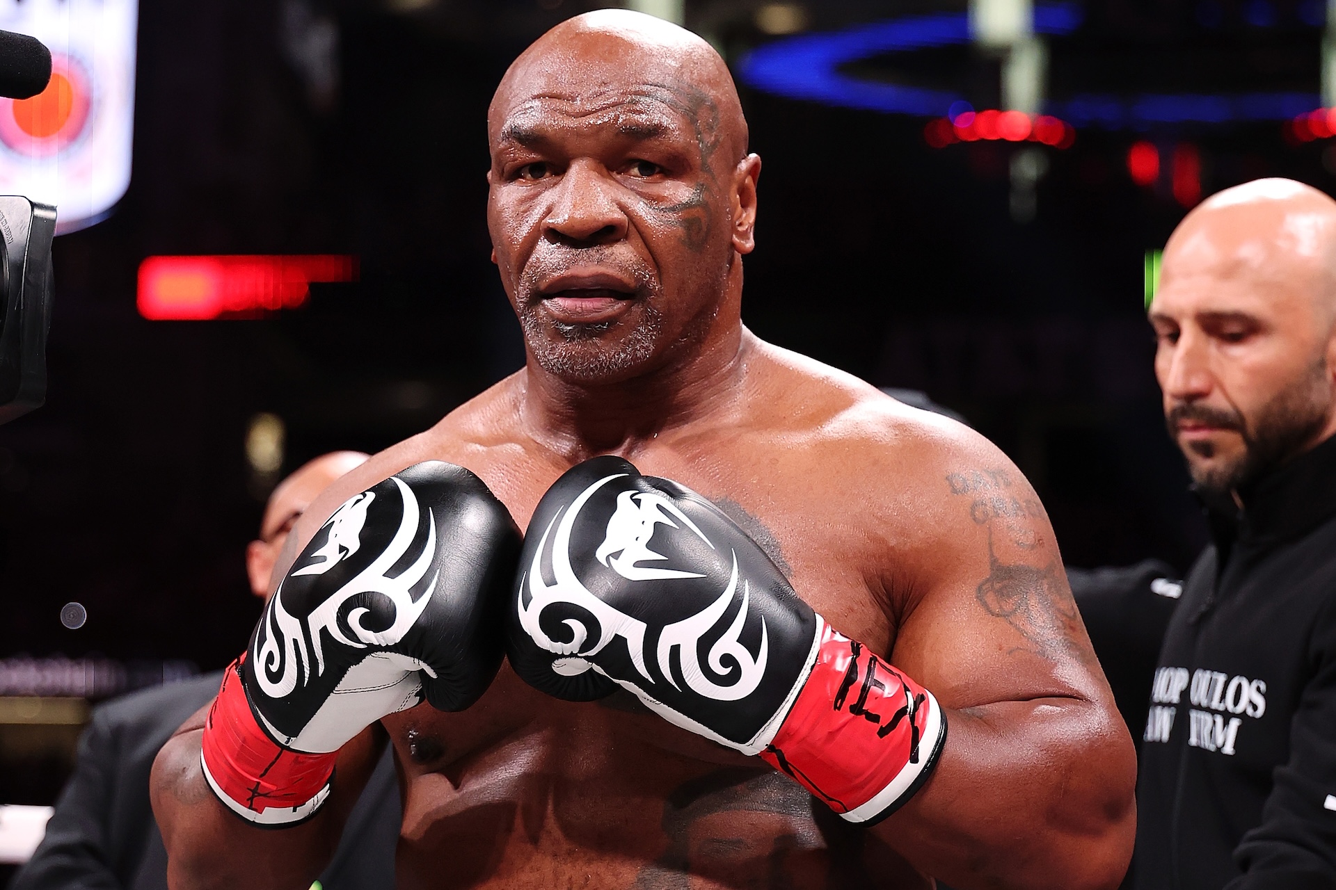 Mike Tyson, shirtless, wearing boxing gloves with a focused expression. A man stands in the background.