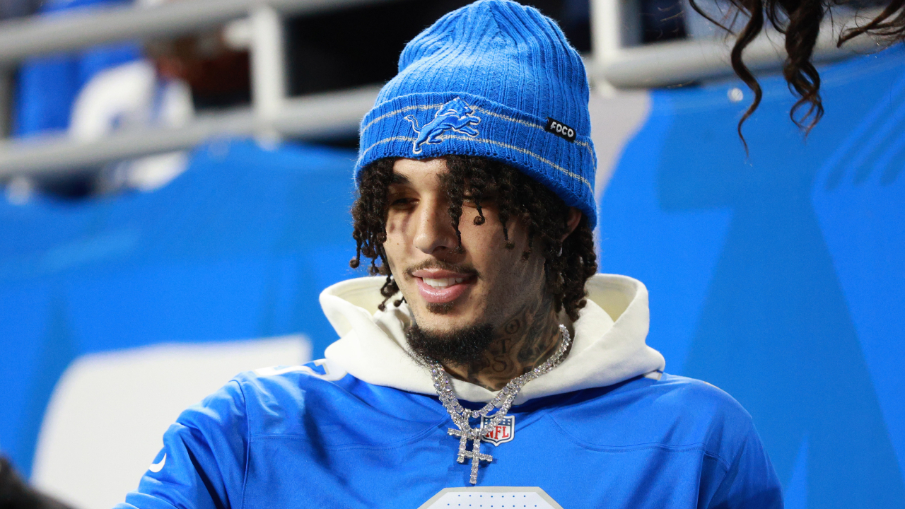 LiAngelo Ball wearing a blue Detroit Lions beanie and jersey, with tattoos and a cross necklace, smiling at an event.