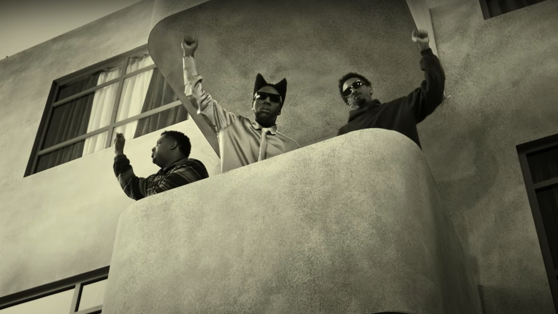Three men on a balcony, raising their fists in a black and white image. They wear sunglasses and look towards the sky.