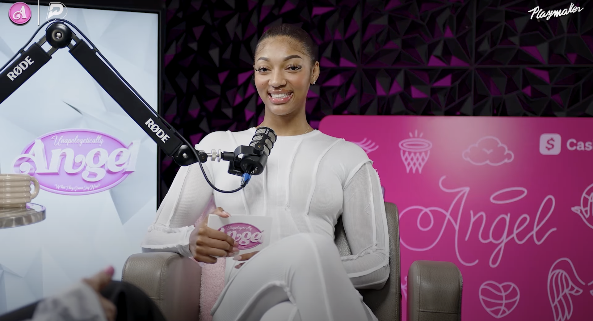 A woman in a white outfit sits in a podcast studio with a microphone. The background has pink signage with "Angel" written on it.