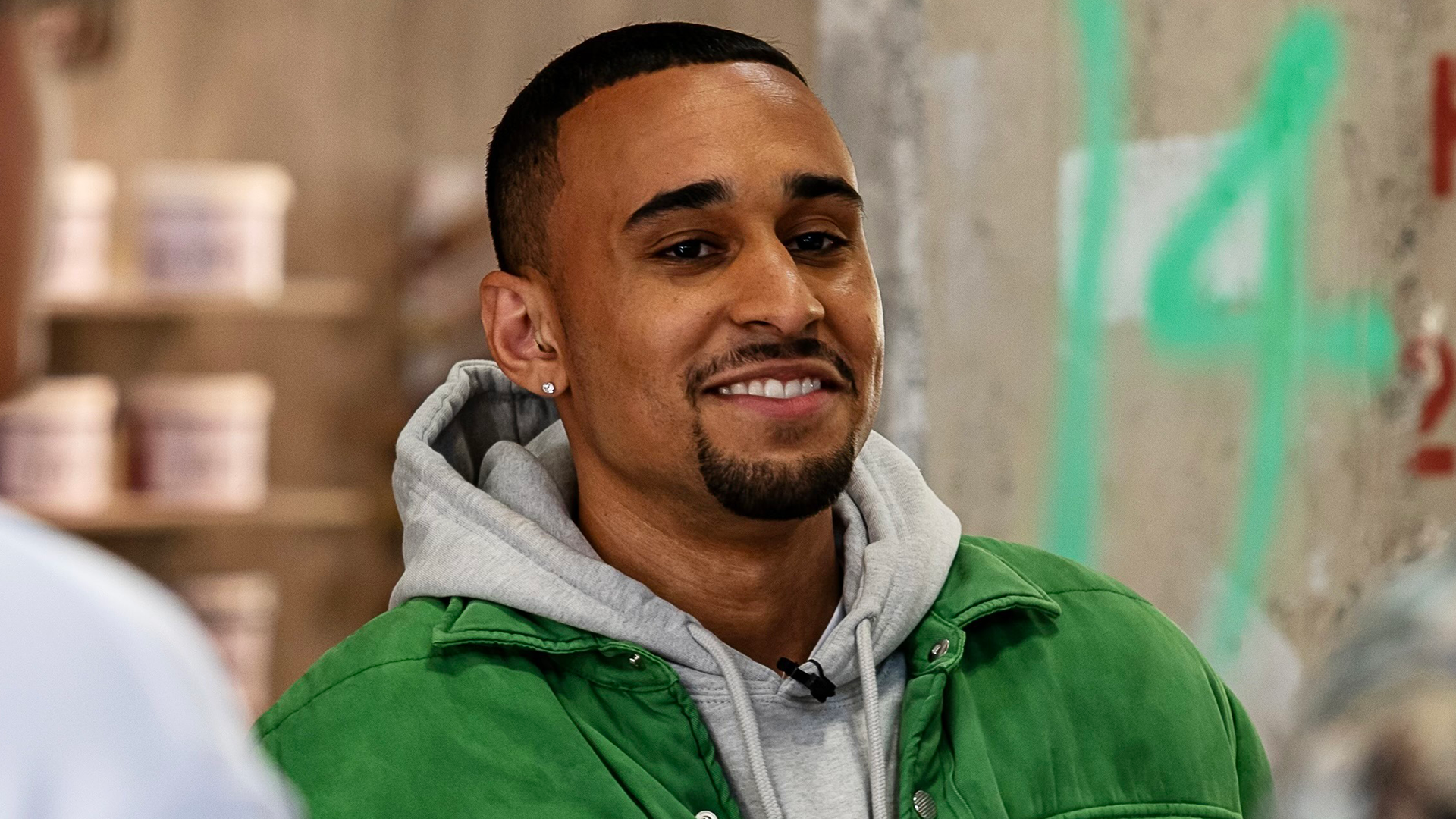 A person smiling, wearing a green jacket over a gray hoodie, standing indoors with a blurred background.
