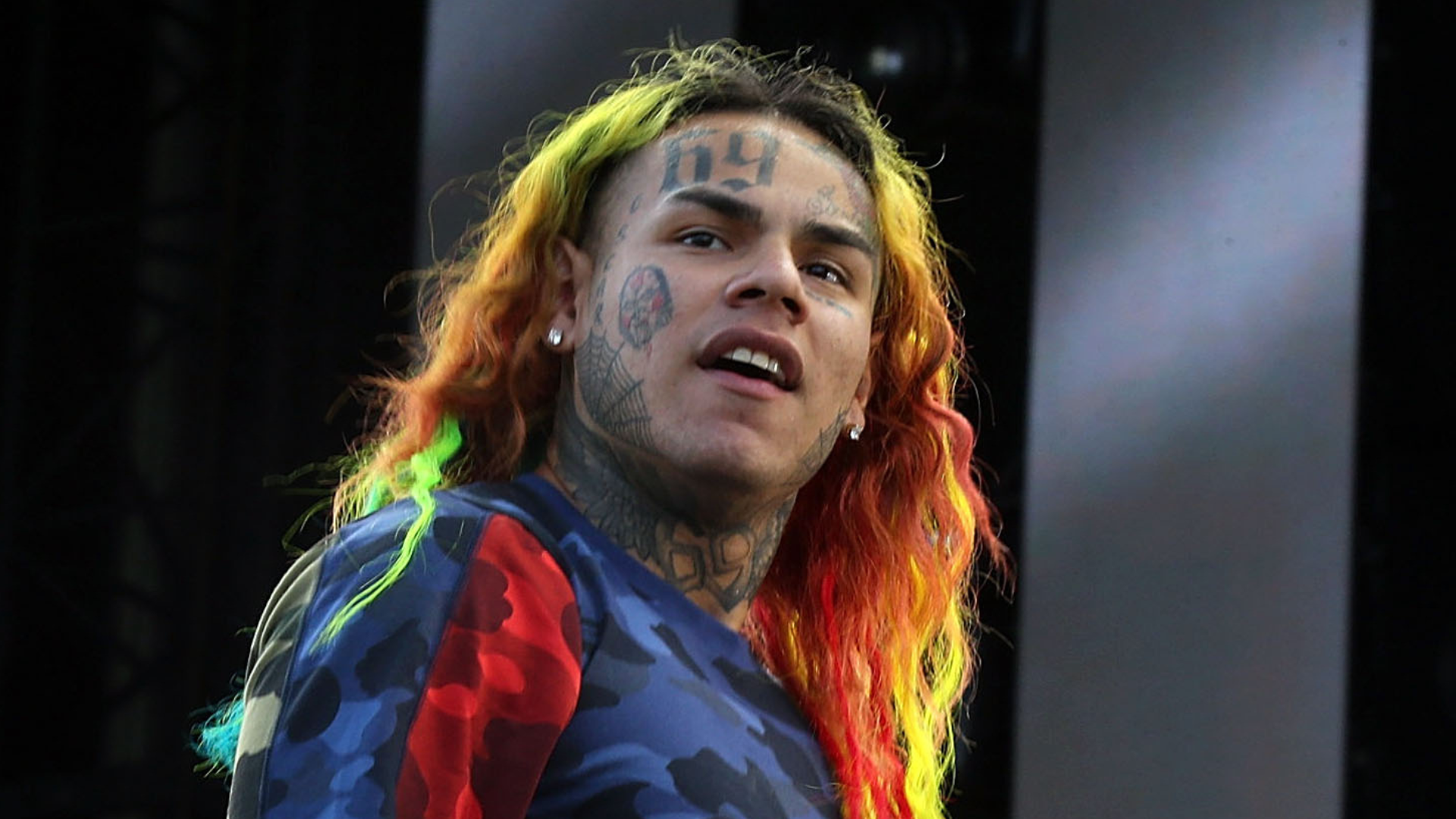 Rapper Tekashi 6ix9ine with colorful hair and facial tattoos, wearing a camo-patterned shirt, performing on stage.