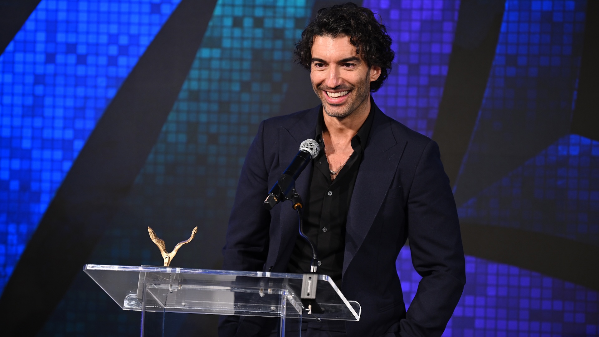 Justin Baldoni standing at a podium with a microphone stand and wearing a blue blazer.
