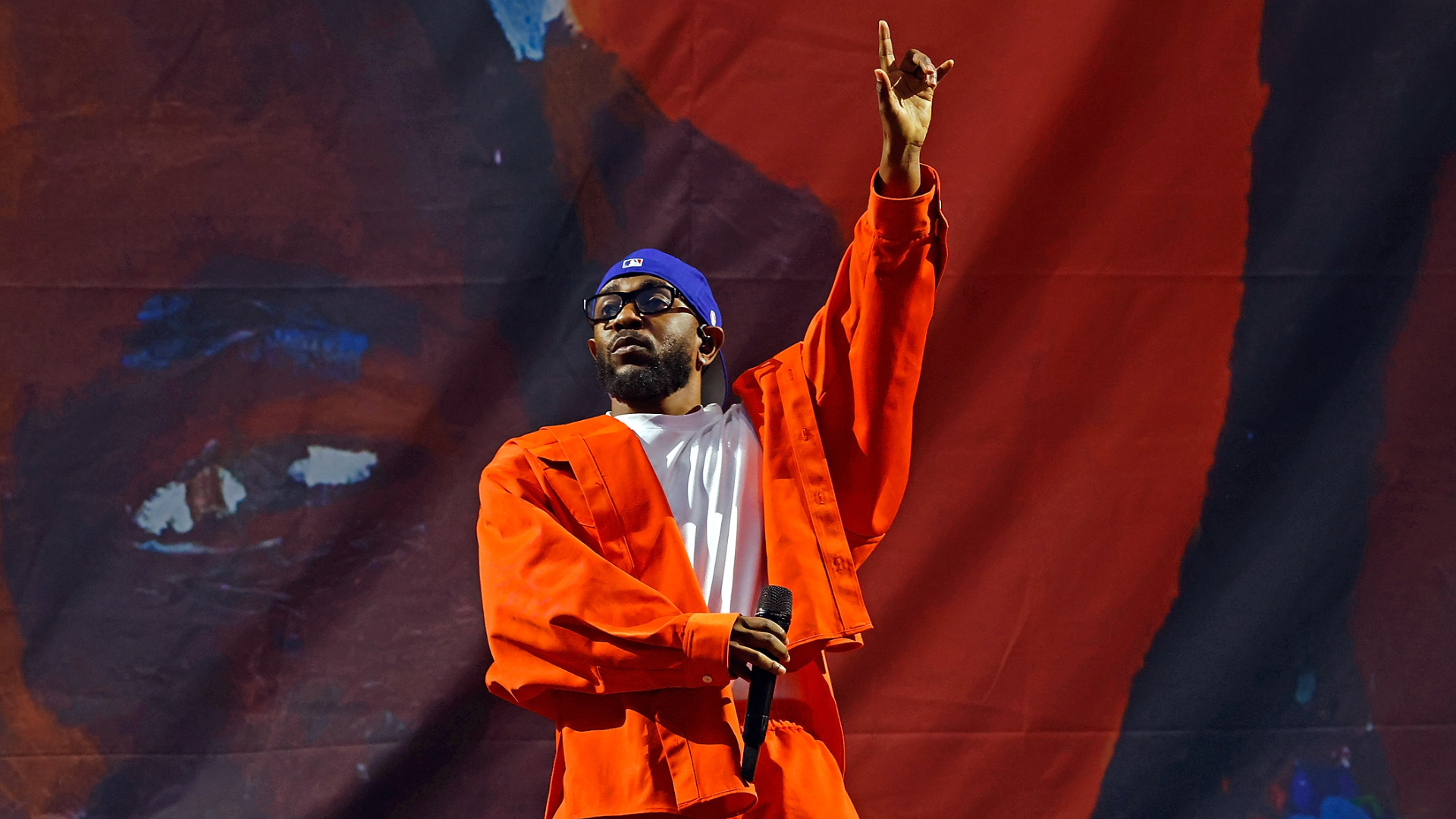Kendrick Lamar in an orange outfit and blue cap holds a microphone and raises one hand on stage with a colorful backdrop.
