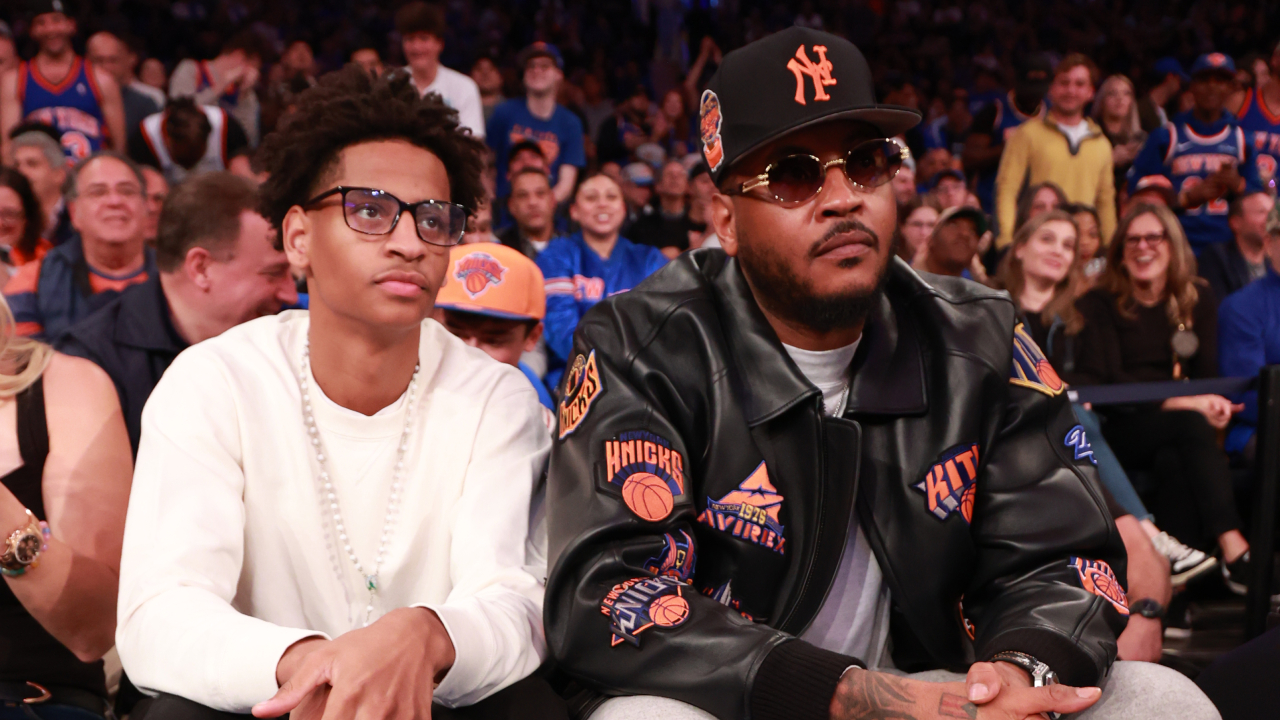 Carmelo Anthony, wearing a Knicks-themed jacket and cap, sits courtside with his son Kiyan at a basketball game.