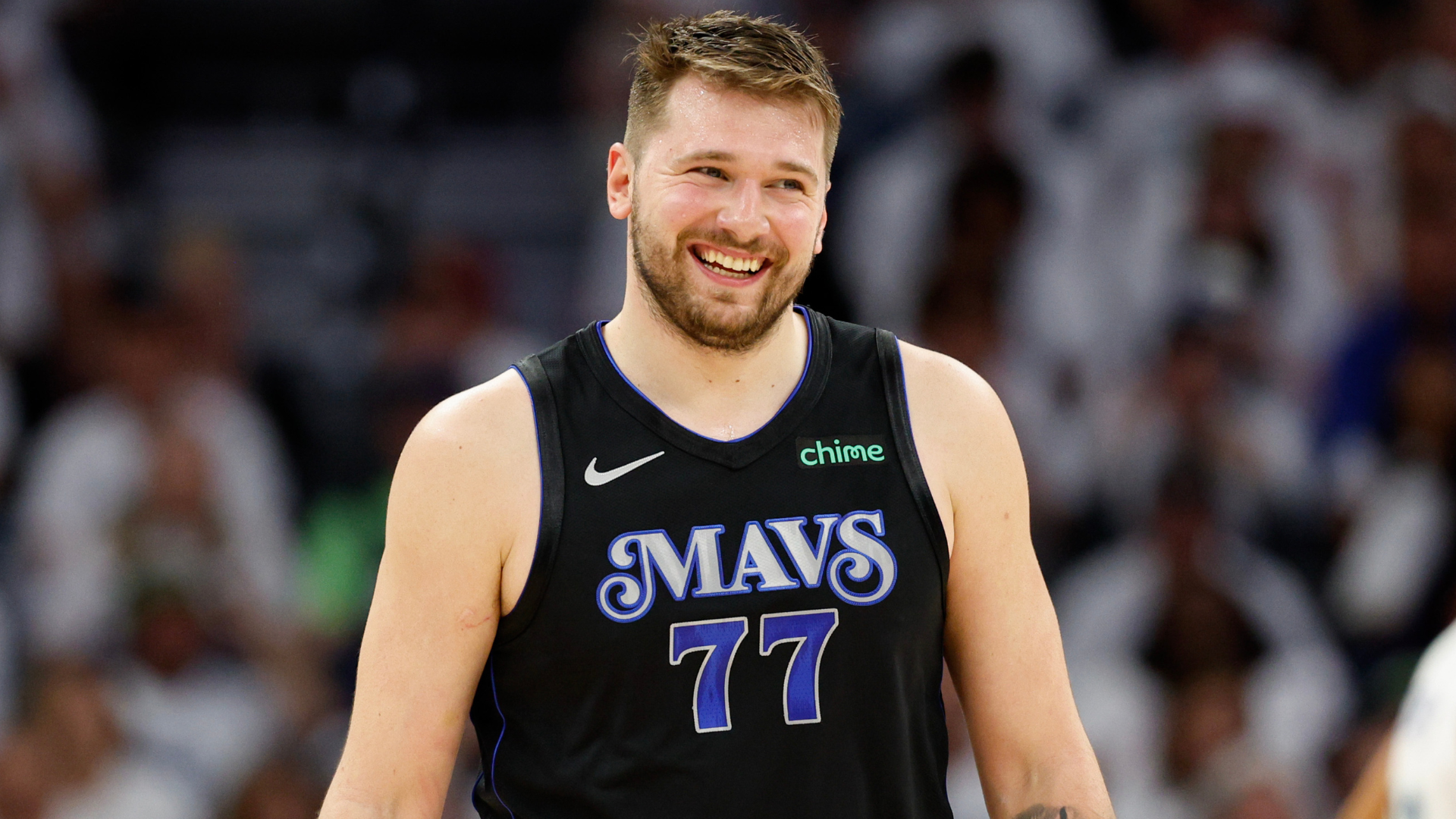 Luka Dončić, wearing a black "Mavs" jersey, smiles on a basketball court.