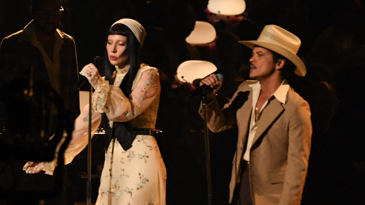 Lady Gaga in a vintage dress and headpiece sings with Bruno Mars in a hat and suit on stage, under warm lighting.