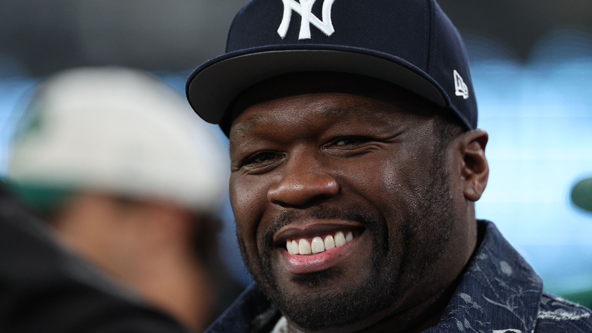 50 Cent smiling, wearing a New York Yankees cap and a patterned jacket.