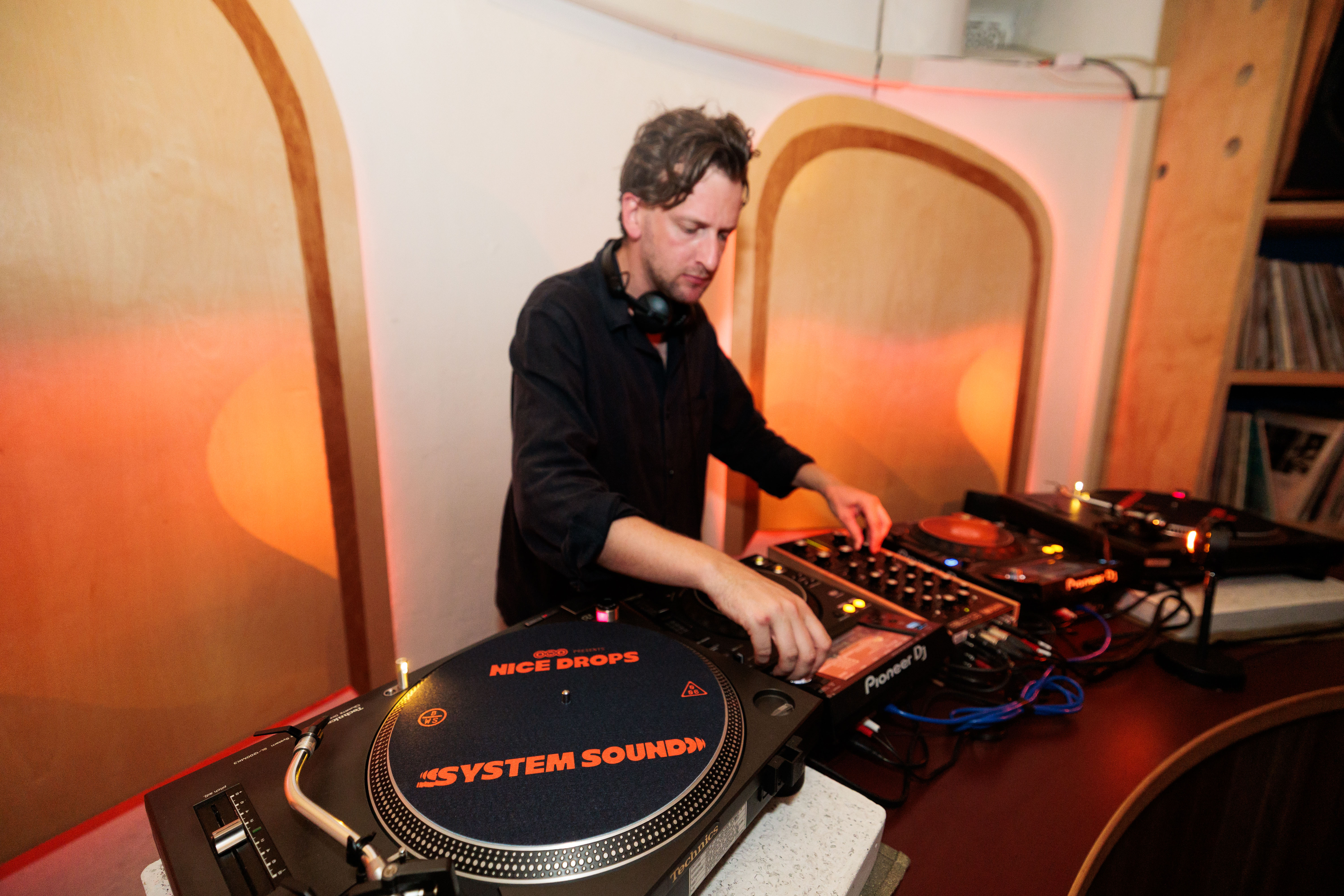 A DJ is mixing music on turntables in a warmly lit room. The equipment includes a turntable with "SYSTEM SOUND" on the slipmat.