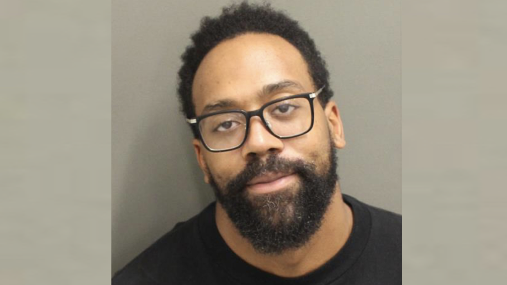 Marcus Jordan with glasses and a beard, wearing a black shirt, against a plain background for police mugshot.
