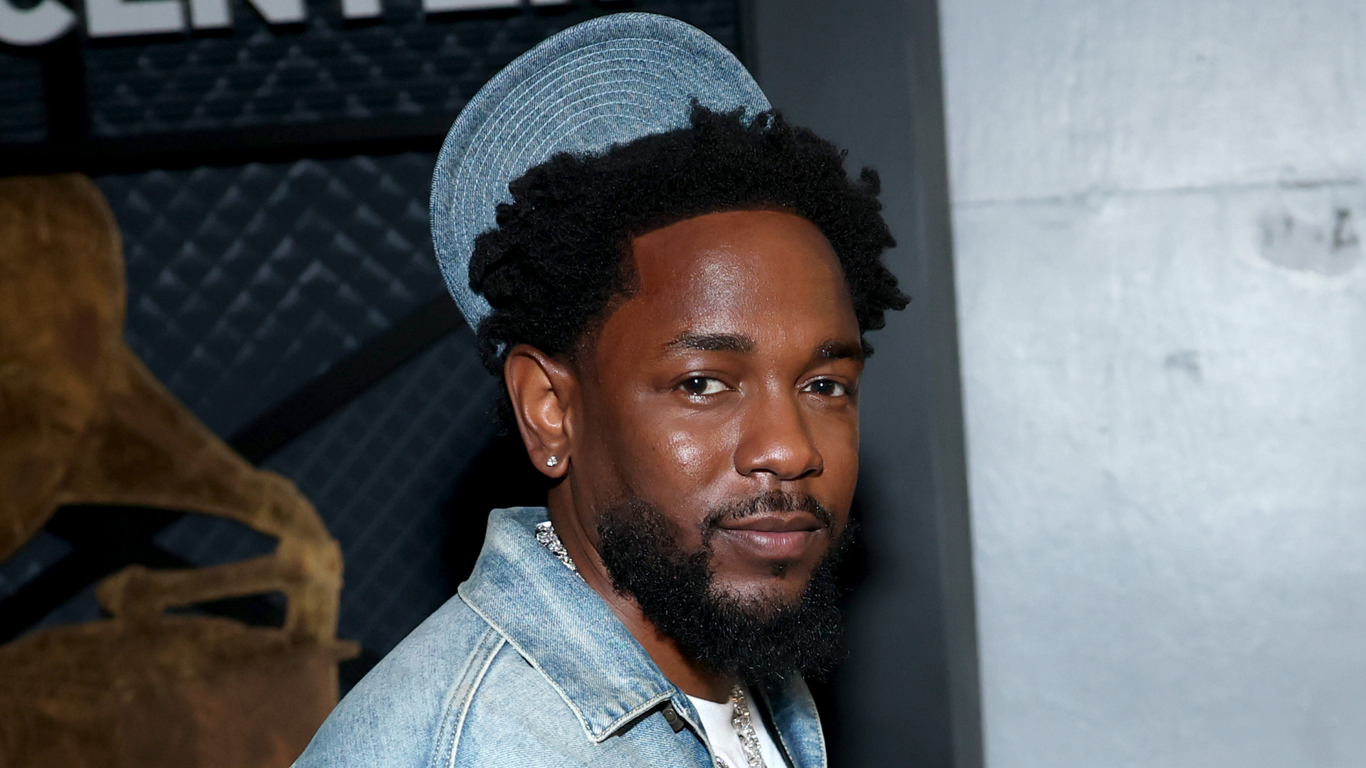 Kendrick Lamar wearing a denim jacket, standing in front of a dark background with a subtle smile.