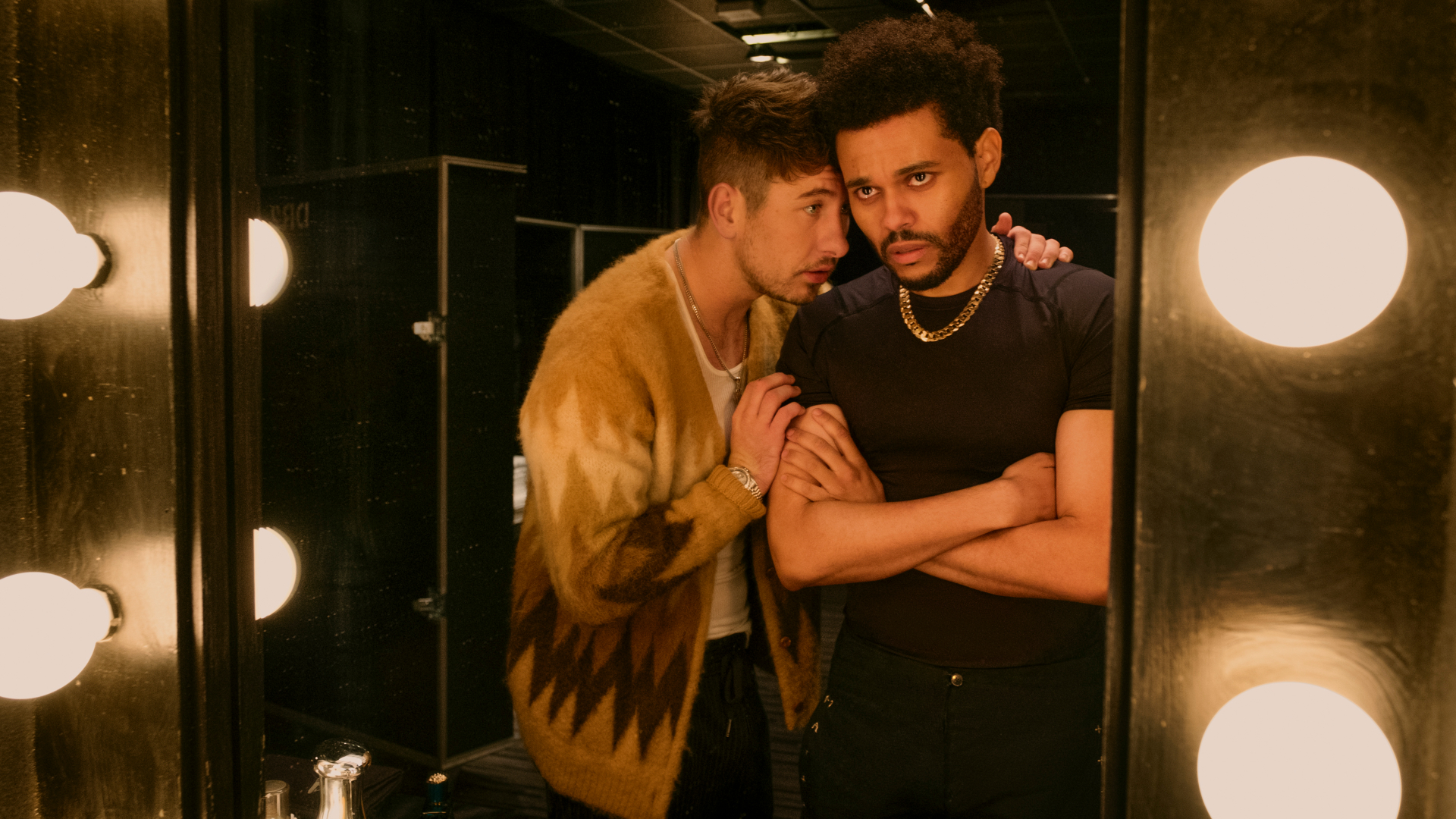 Two men in a dimly lit backstage area, with one whispering to the other. Bright vanity lights frame the scene.