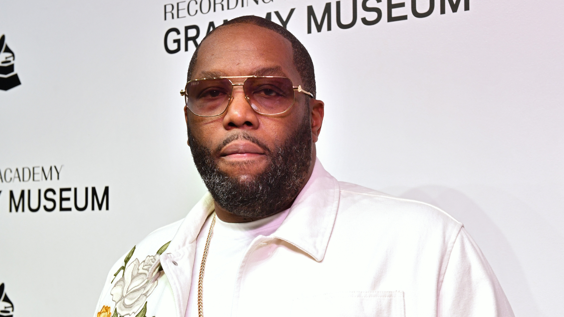 Killer Mike at the Grammy Museum, wearing a white jacket and sunglasses, standing against a backdrop with Grammy Museum text.