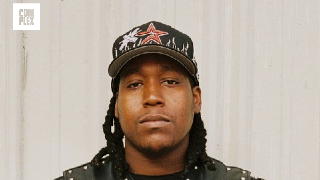 Don Toliver wearing a black cap with a star design and a studded jacket, standing in front of a light-colored wall.