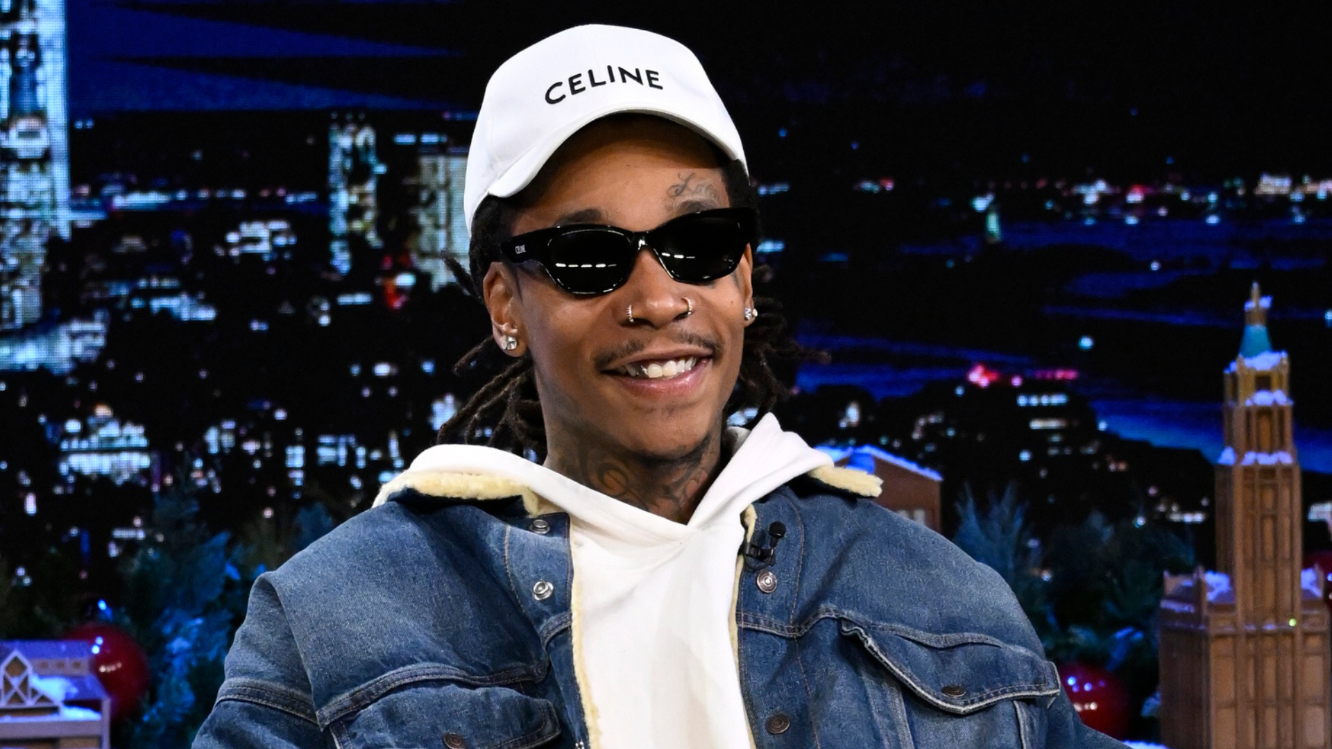 A person wearing a white Celine cap, sunglasses, a denim jacket, and a white hoodie, sitting in a nighttime cityscape setting.