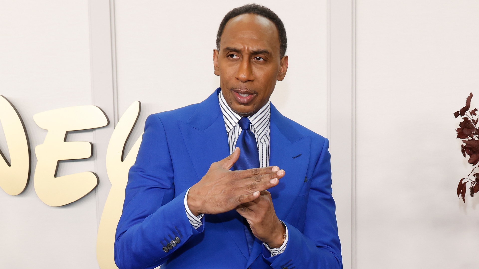Stephen A. Smith in a bright blue suit gestures with his hands in front of a neutral backdrop.