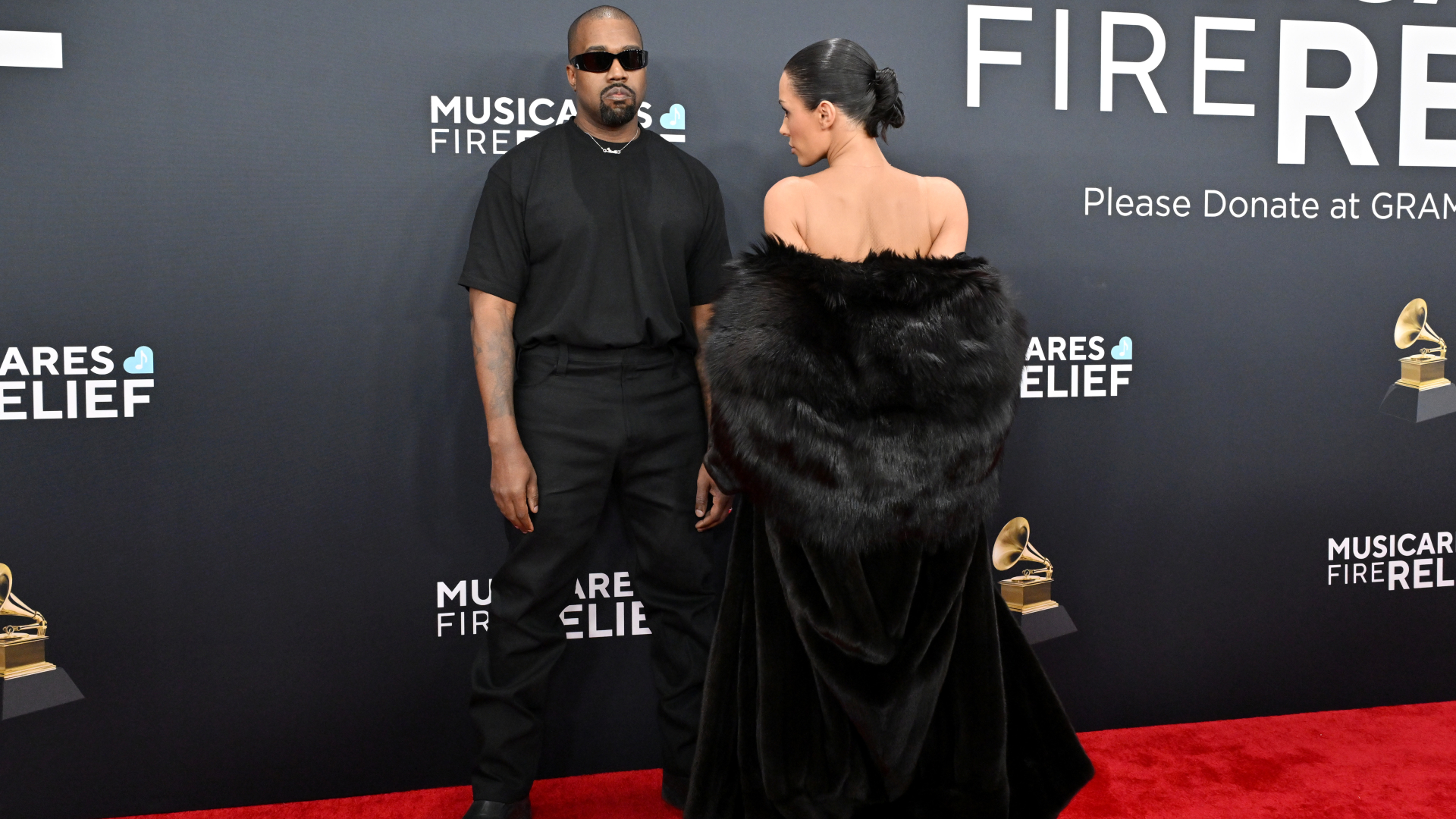 Ye in sunglasses and black outfit stands on the red carpet with Bianca Censori in a black fur coat