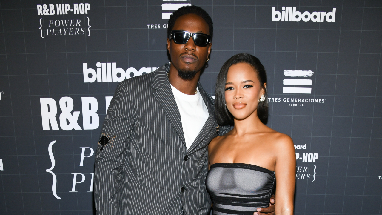 Serayah on a runway wearing a red lingerie set and a pink tulle robe, smiling and holding her pregnant belly.