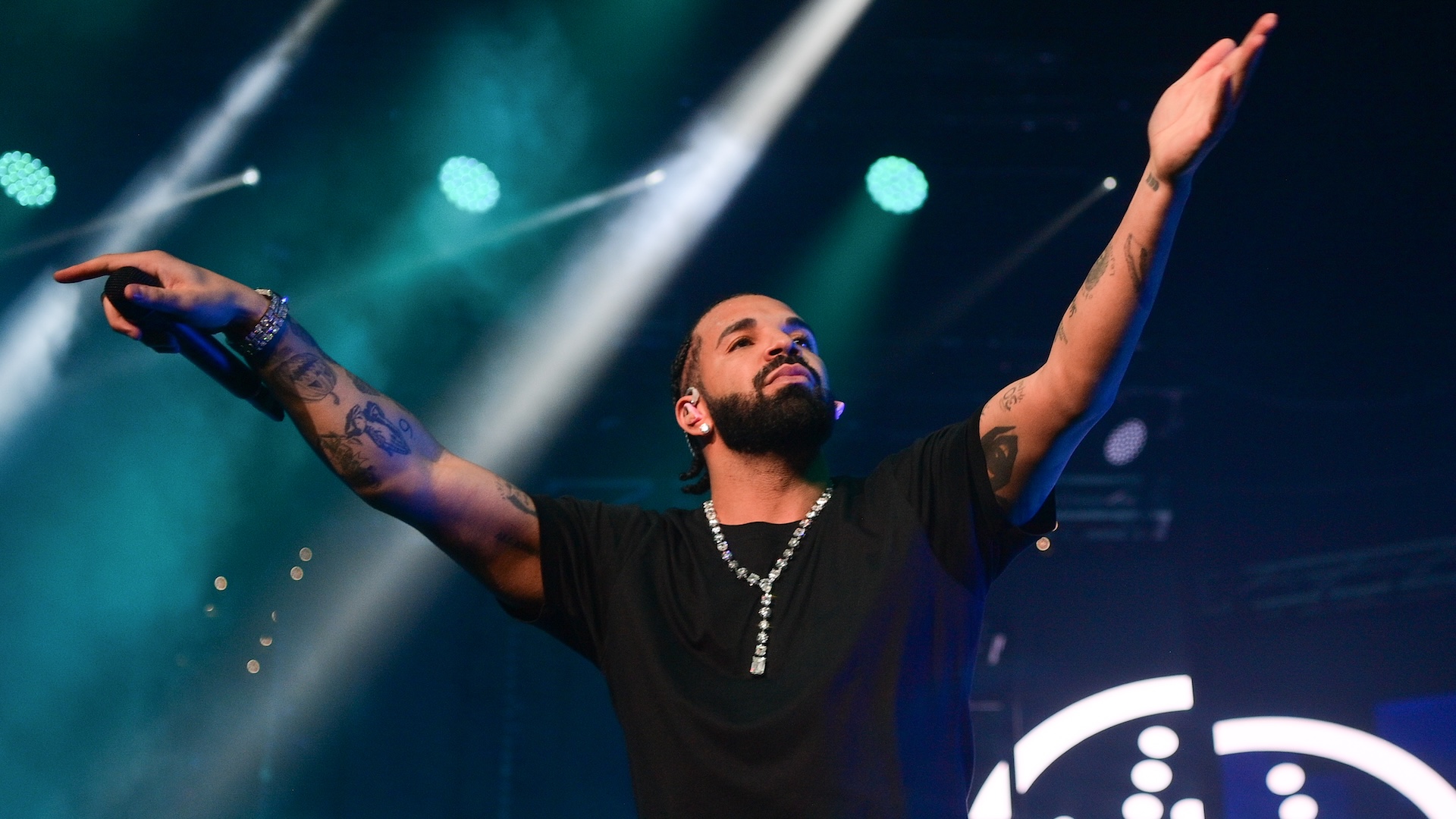 Drake performing on stage with arms raised, wearing a black shirt and necklace, under colorful stage lights.