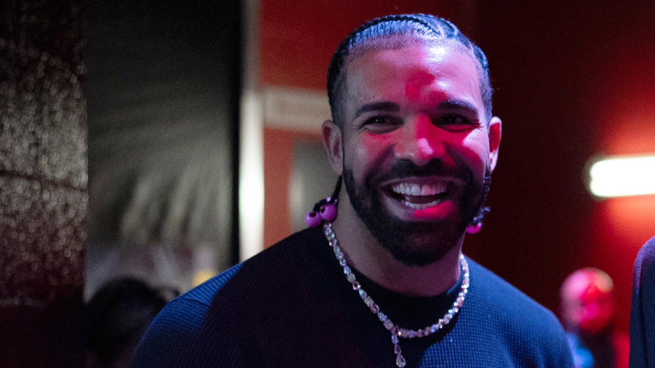 Drake smiling, wearing a black shirt and beaded necklace, with braided hair. The background is dimly lit with red and blue hues.