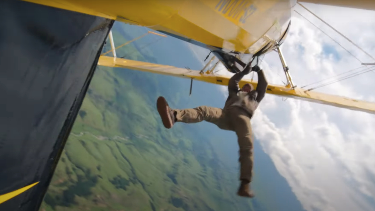 A person hanging from the wing of a yellow biplane in mid-air, with a landscape of green fields and clouds in the background.