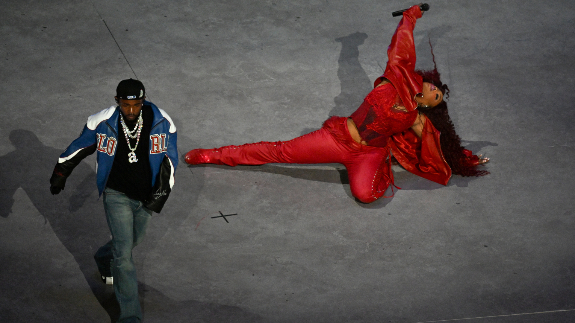 A performer in a red outfit is energetically singing on stage, while another person in a blue jacket walks nearby.