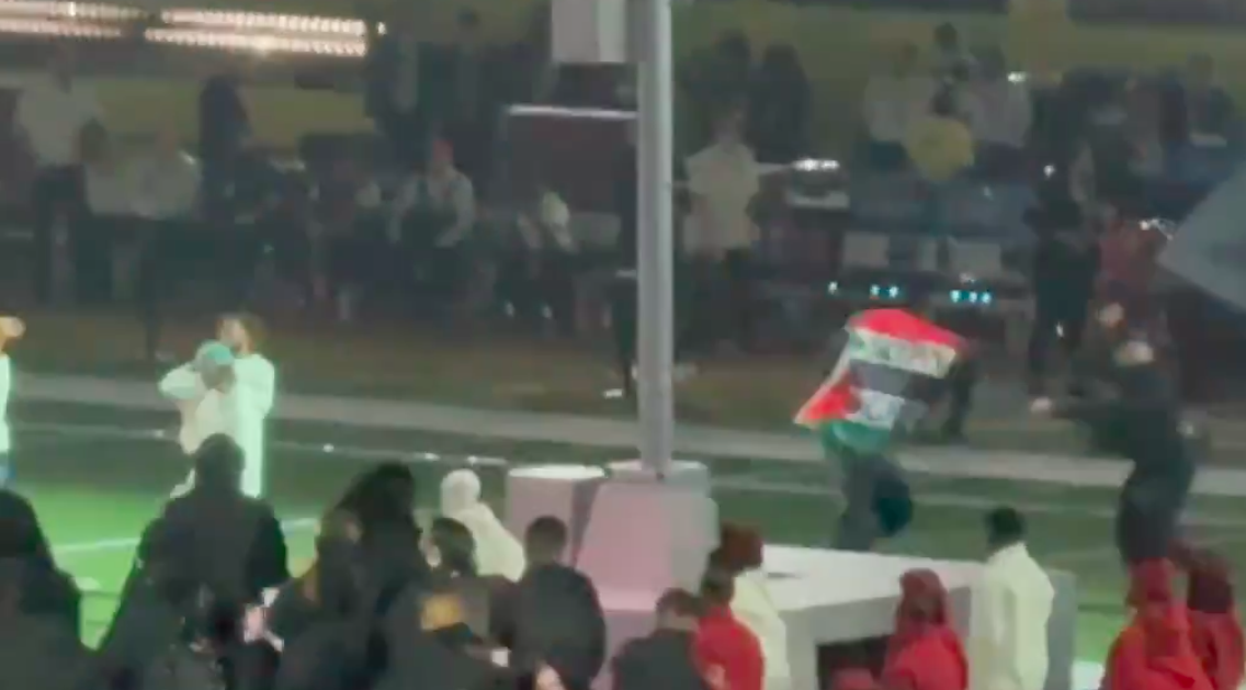 A person runs on a field holding an Iraqi flag. People are standing around, and the scene appears to be at a sports event or concert.