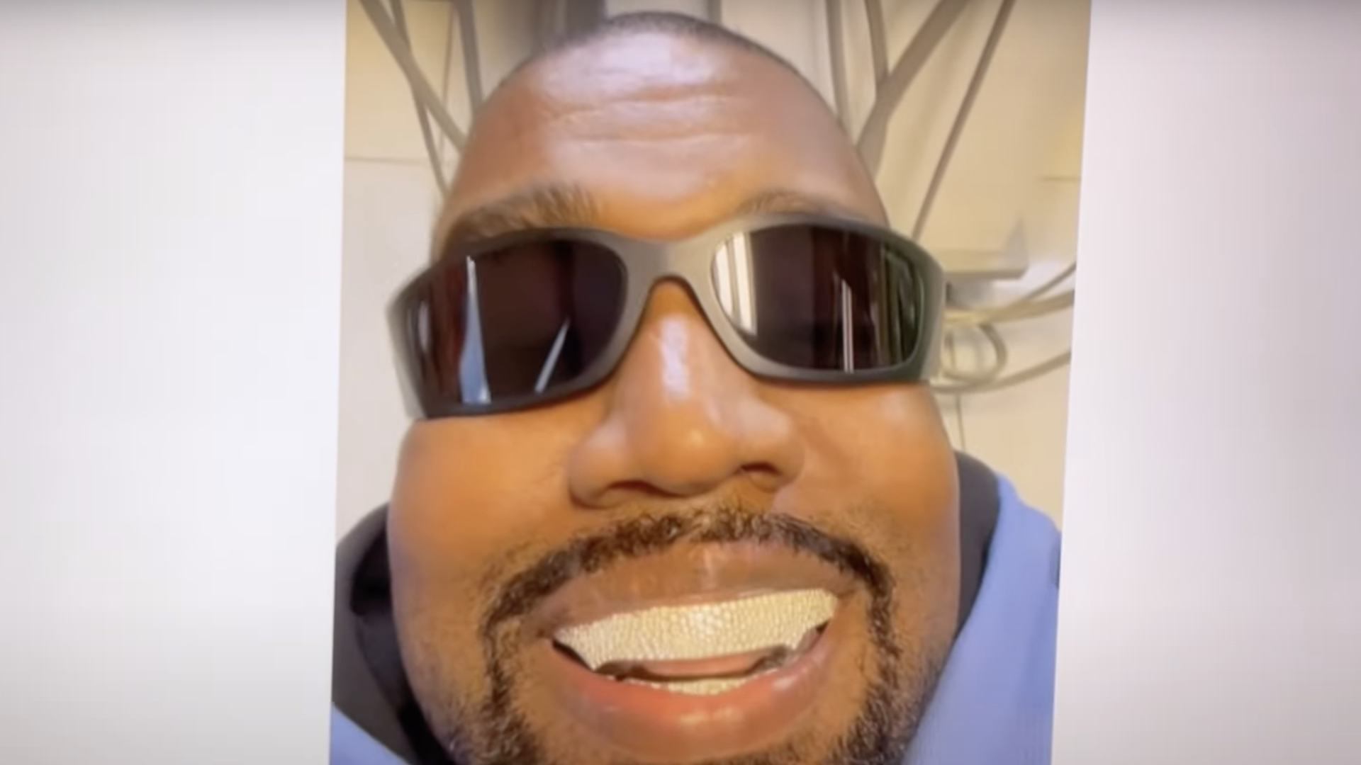 A person wearing sunglasses and a hoodie smiles with a grill on their teeth.