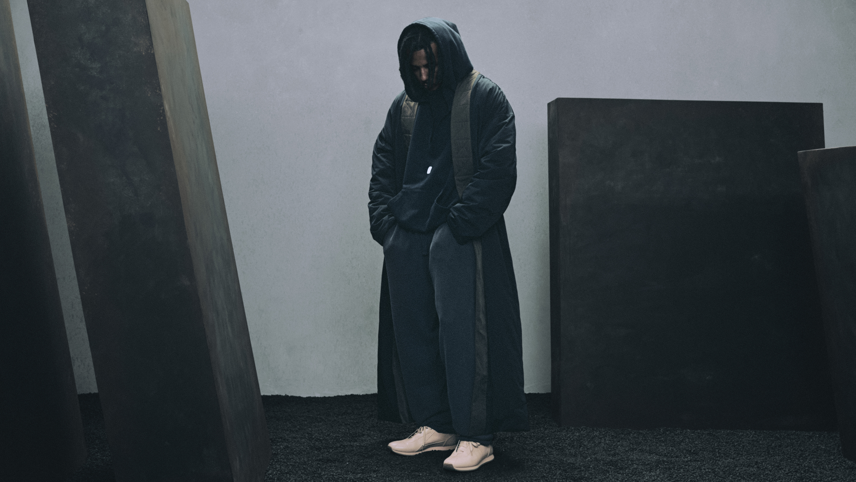 A person in a hooded coat stands among large dark rectangular structures, hands in pockets, wearing light-colored sneakers.