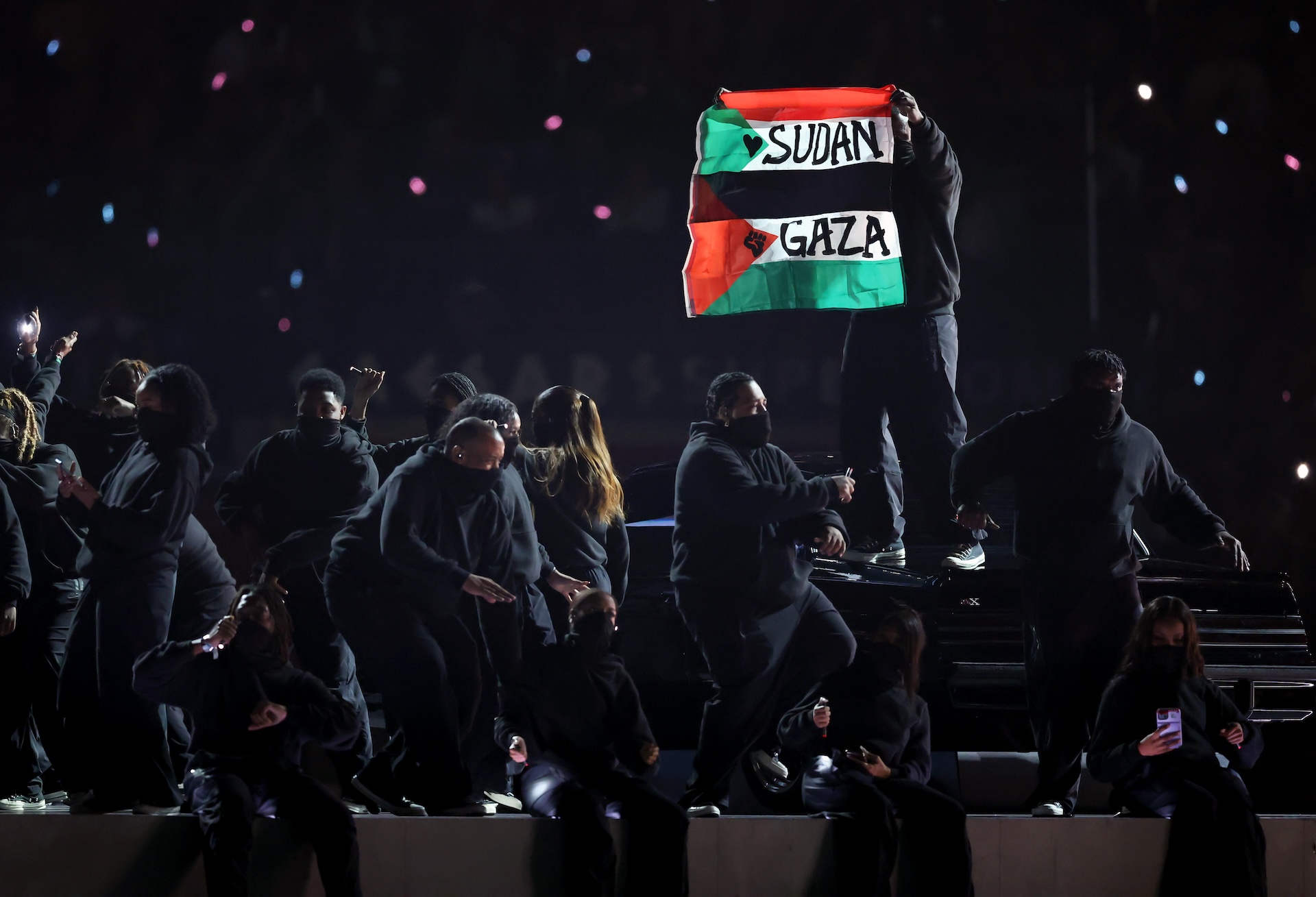 Performers on stage, one holding a flag with "Sudan" and "Gaza" written on it. The scene is dimly lit with a dark backdrop.