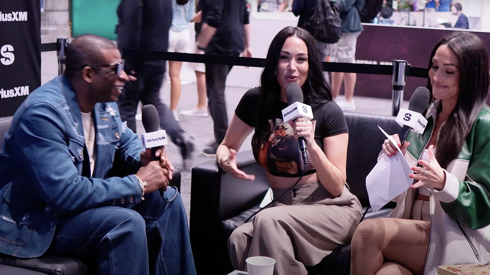 Deion Sanders in a denim jacket is being interviewed by Nikki And Brie, who are holding microphones, sitting on a black couch at a public event.