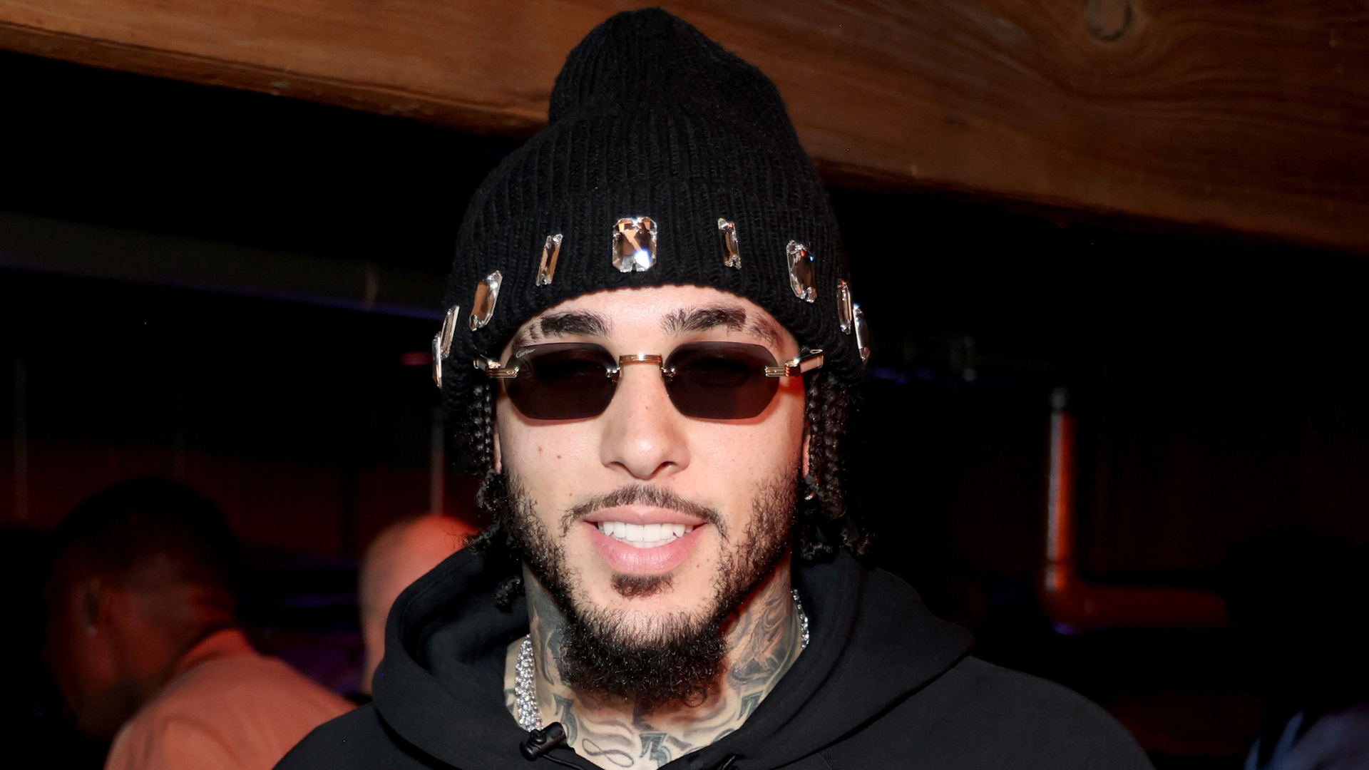 A man wearing sunglasses, a black beanie, and a hoodie, smiling at an indoor event.