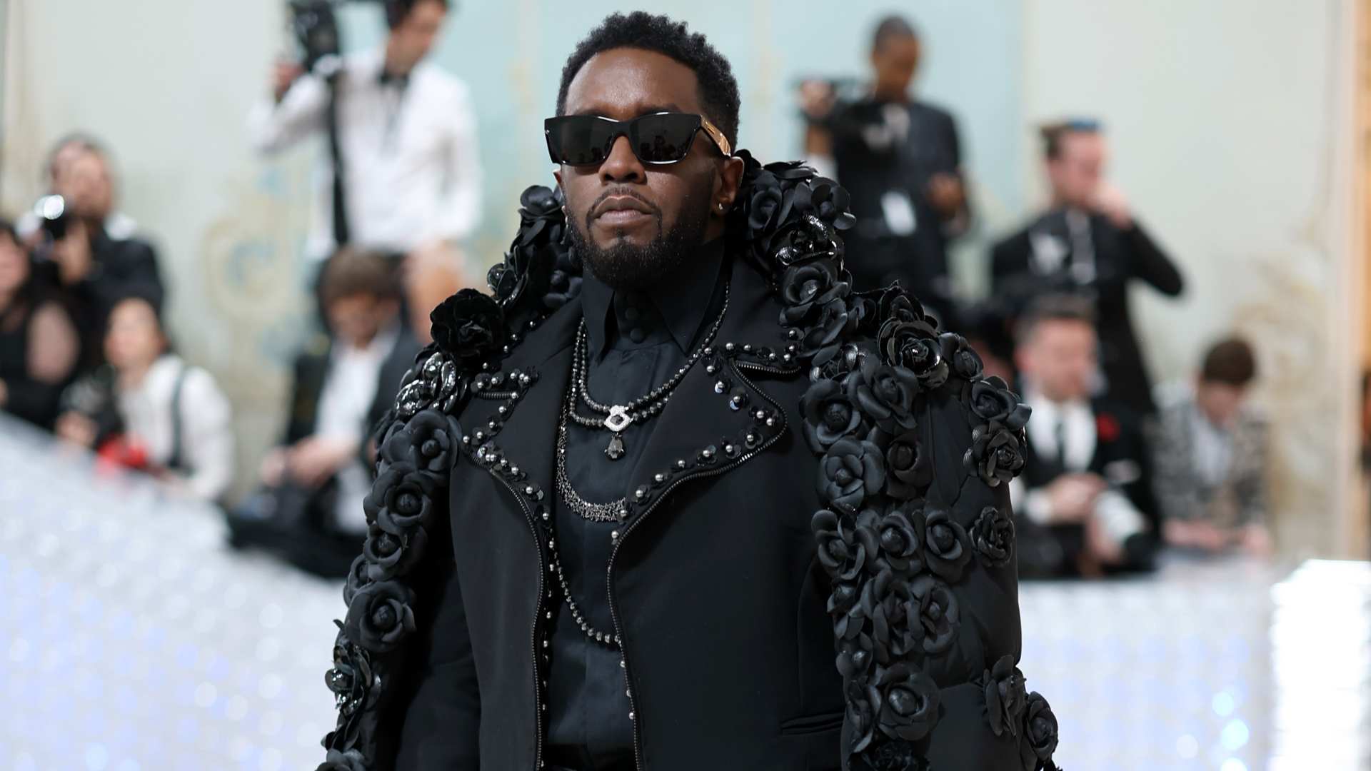 A man in a black outfit adorned with floral details and sunglasses at a formal event, surrounded by people in the background.