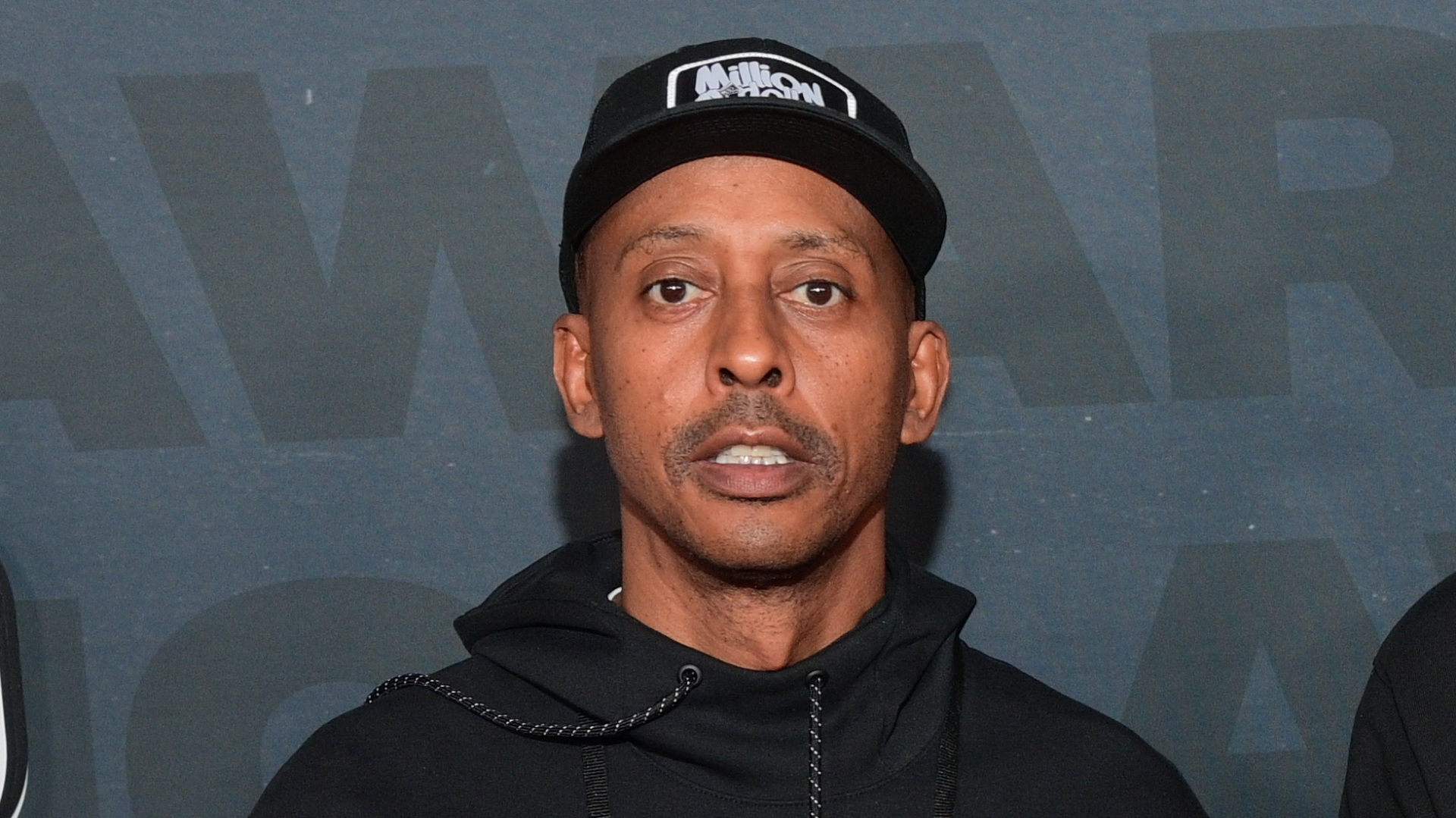 Gillie Da Kid wearing a black hoodie and cap stands against a dark background.